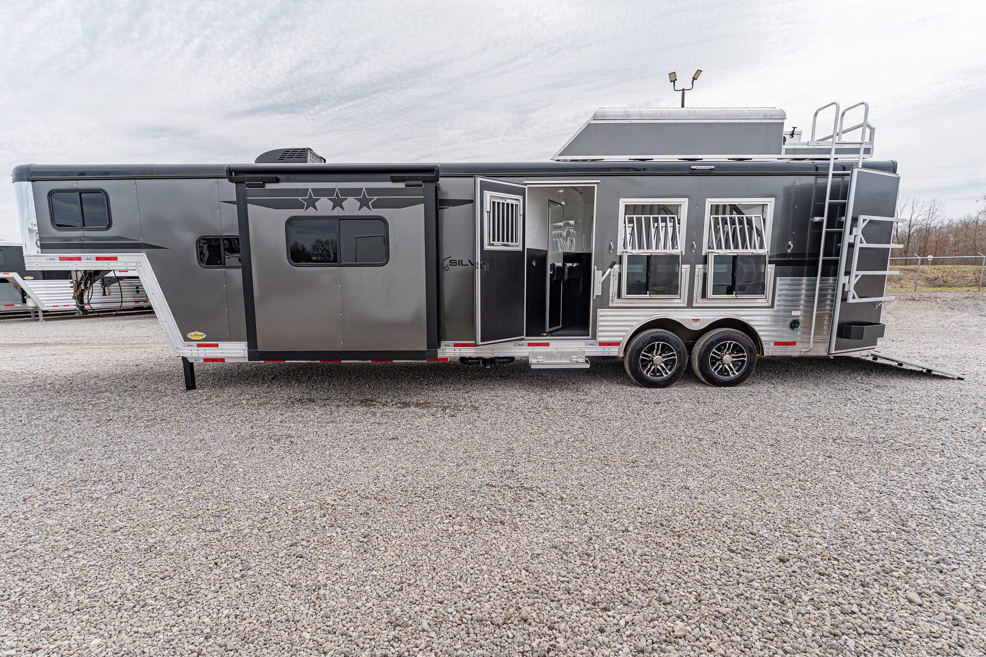 Grey travel trailer with multiple windows parked on a gravel lot; side door open, small stairs extended, and wheel wells visible.
