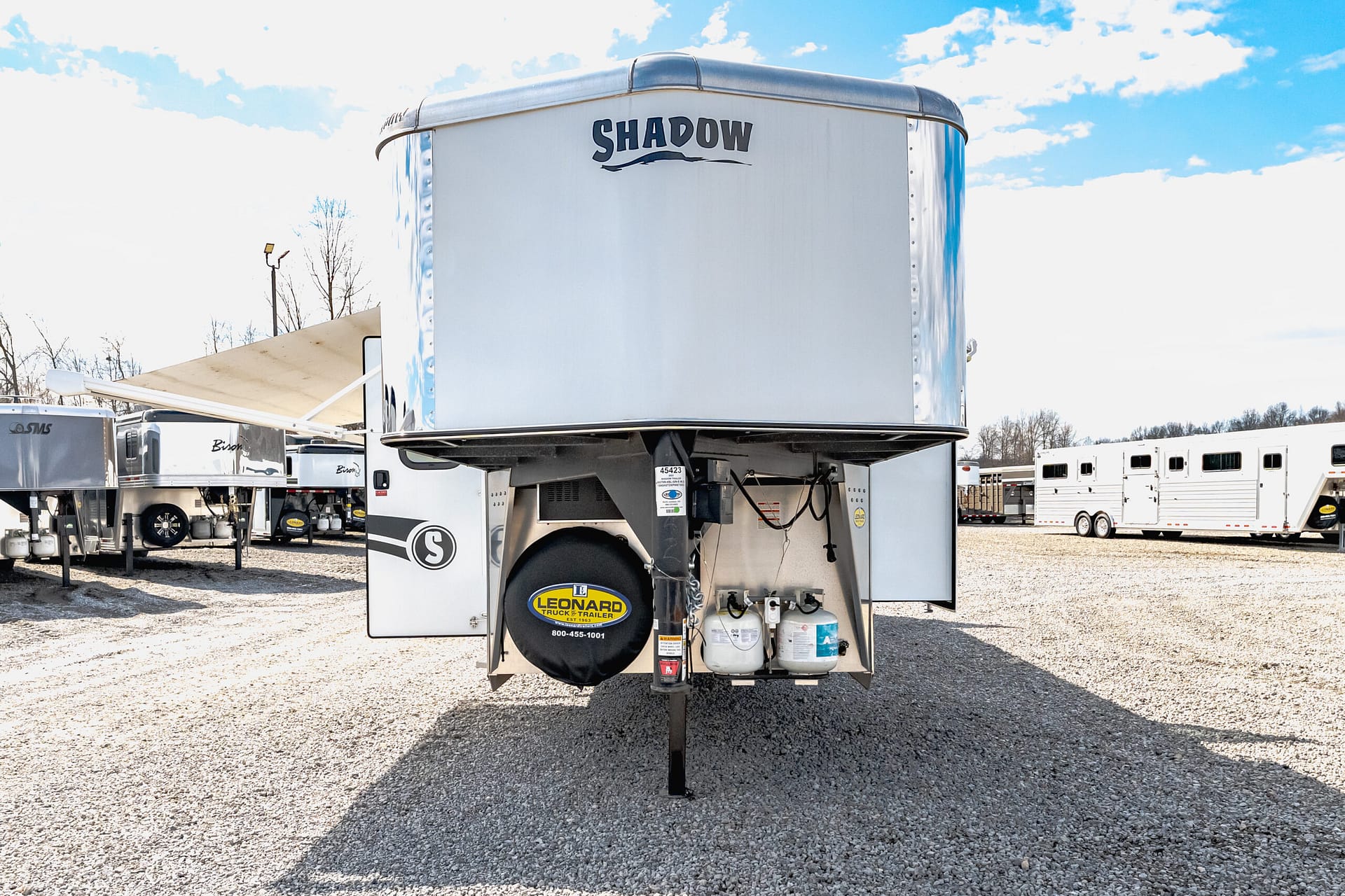 Elevated aluminum trailer on a truck frame in a gravel lot, with a spare tire cover reading Leonard Truck & Trailer and propane tanks underneath.