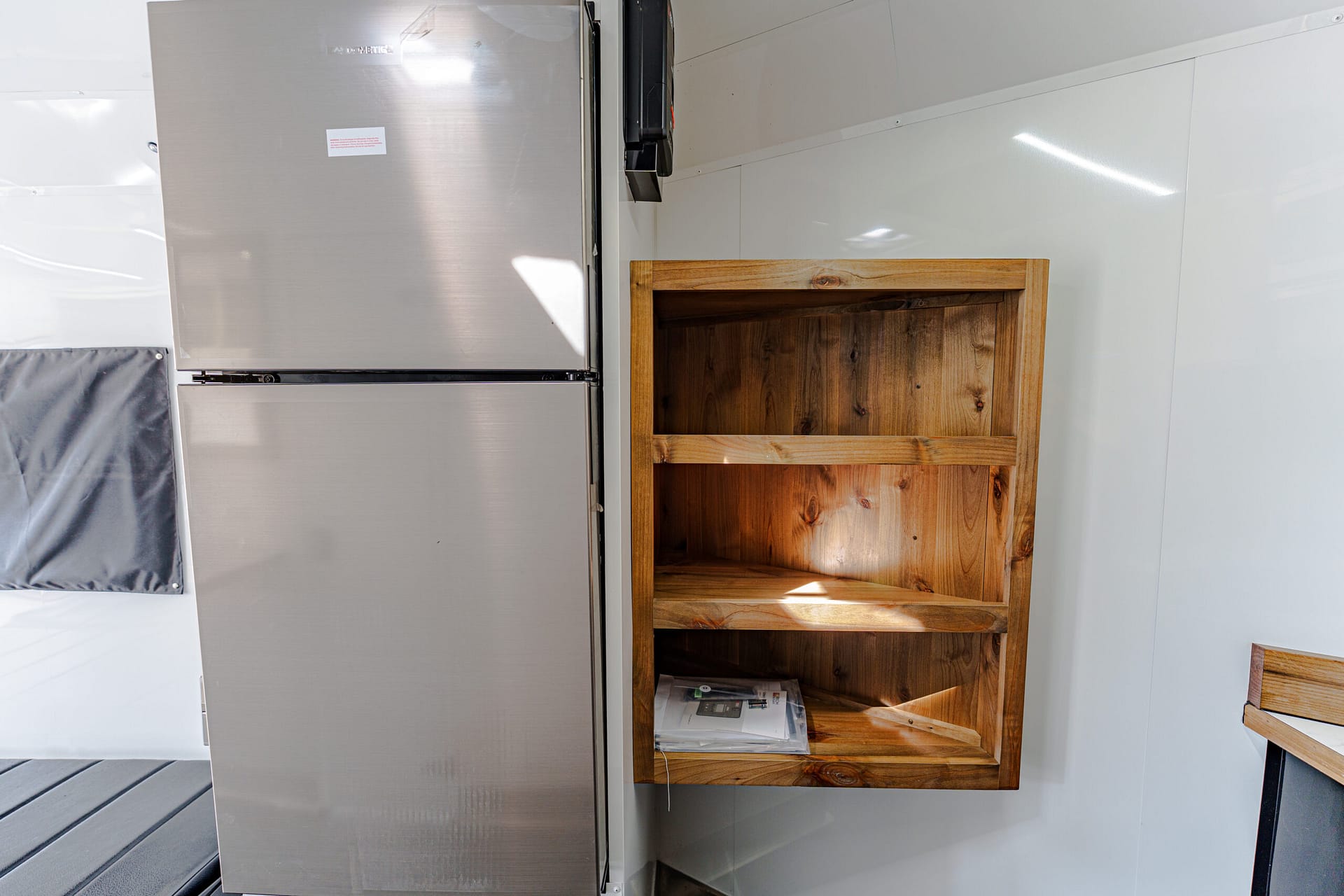Stainless steel refrigerator next to an empty wooden open shelving unit mounted on a white wall in a bright kitchen-like space.