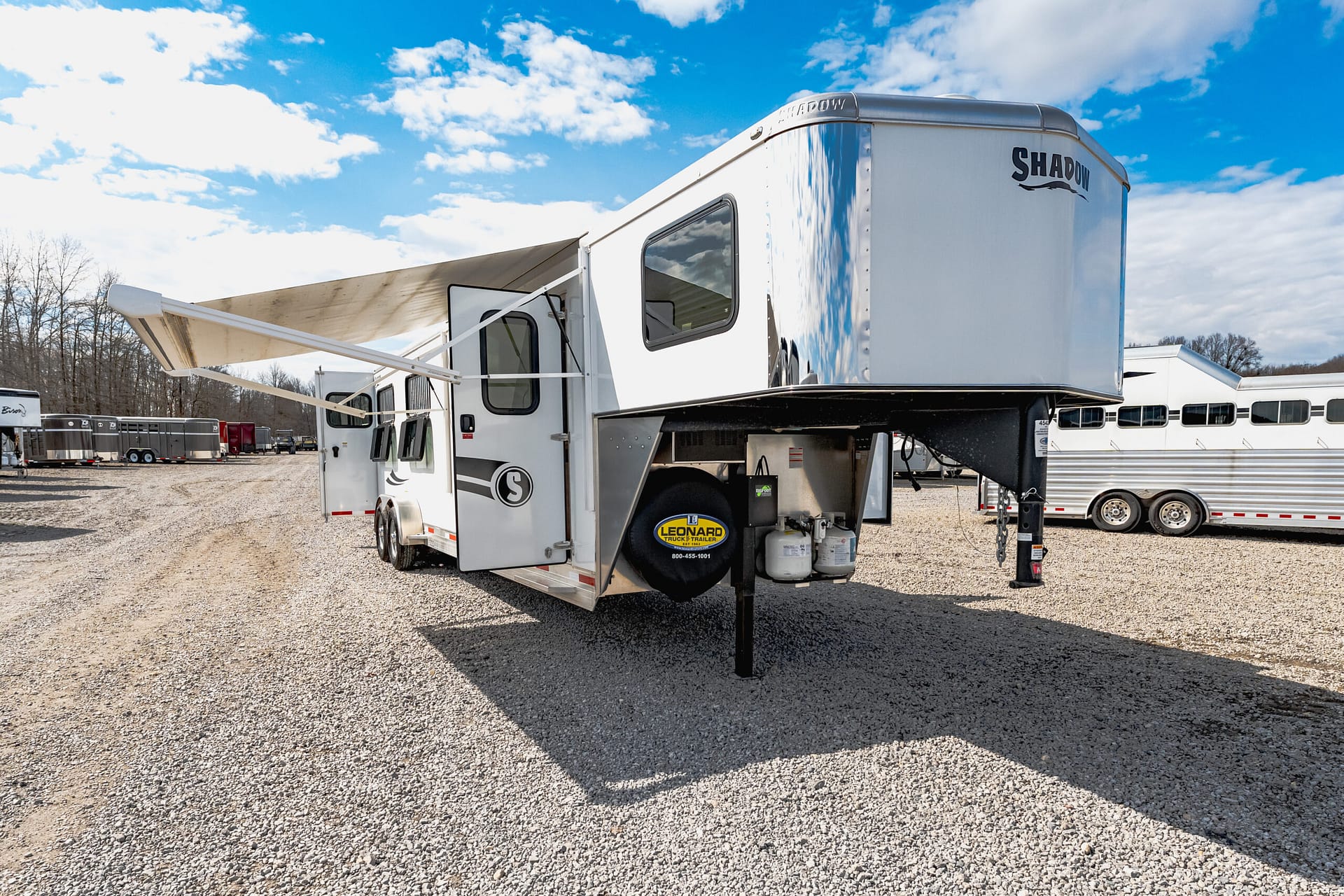 White fifth-wheel RV with extended beige awning on a gravel lot; other trailers visible in the background under a blue sky with clouds.