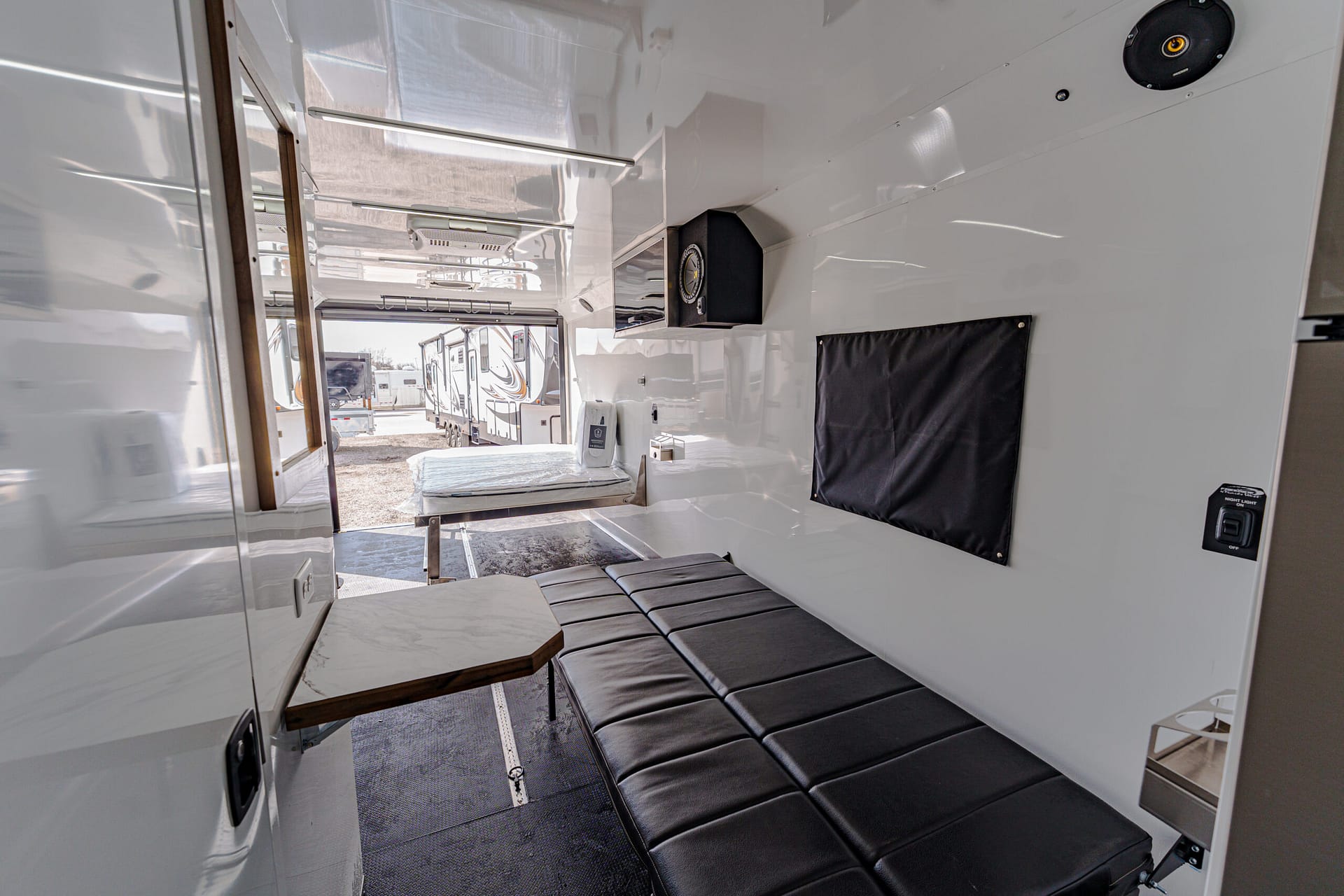 Bright white RV interior with black cushioned bench seating and a small table, view of parked trailers through an open doorway in the background.