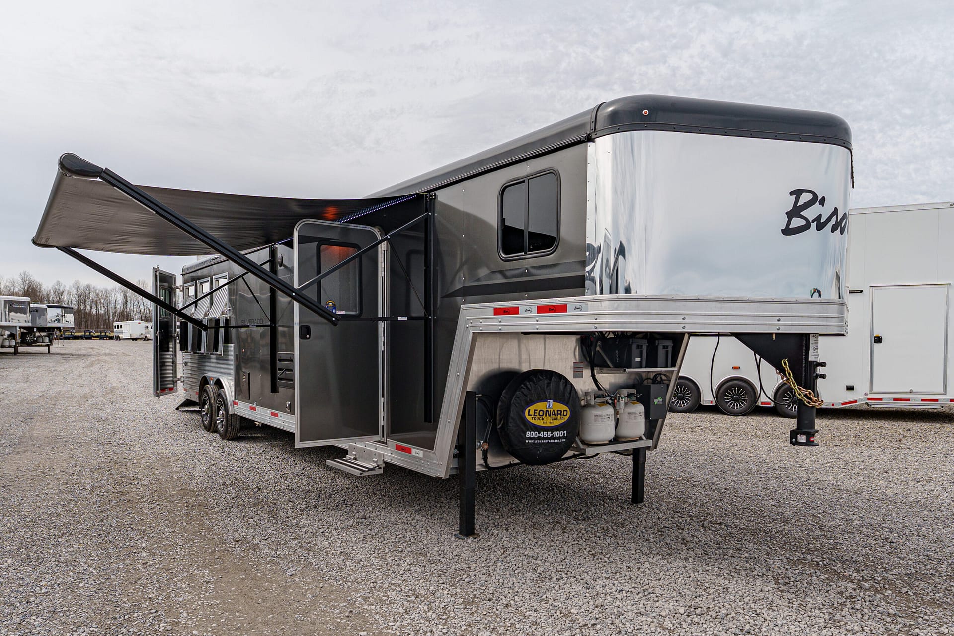 Grey recreational trailer with a large retractable awning extended over steps in a gravel lot; other trailers visible in background.