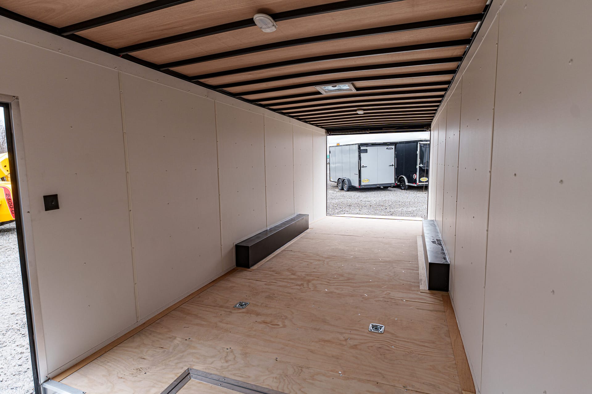 Interior of an empty trailer with a plywood floor and white walls, open to a gravel lot with other trailers outside.