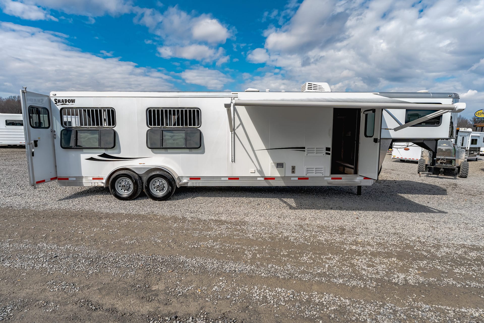 White travel trailer with a retracted awning, parked on a gravel lot under a blue sky with clouds.