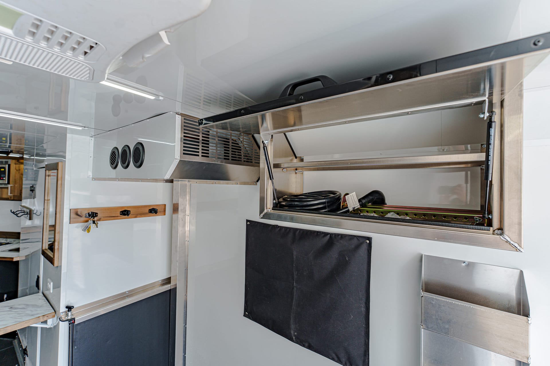 Industrial kitchen trailer interior showing stainless steel equipment, vents, and black fabric panels with hoses and cables stacked overhead.