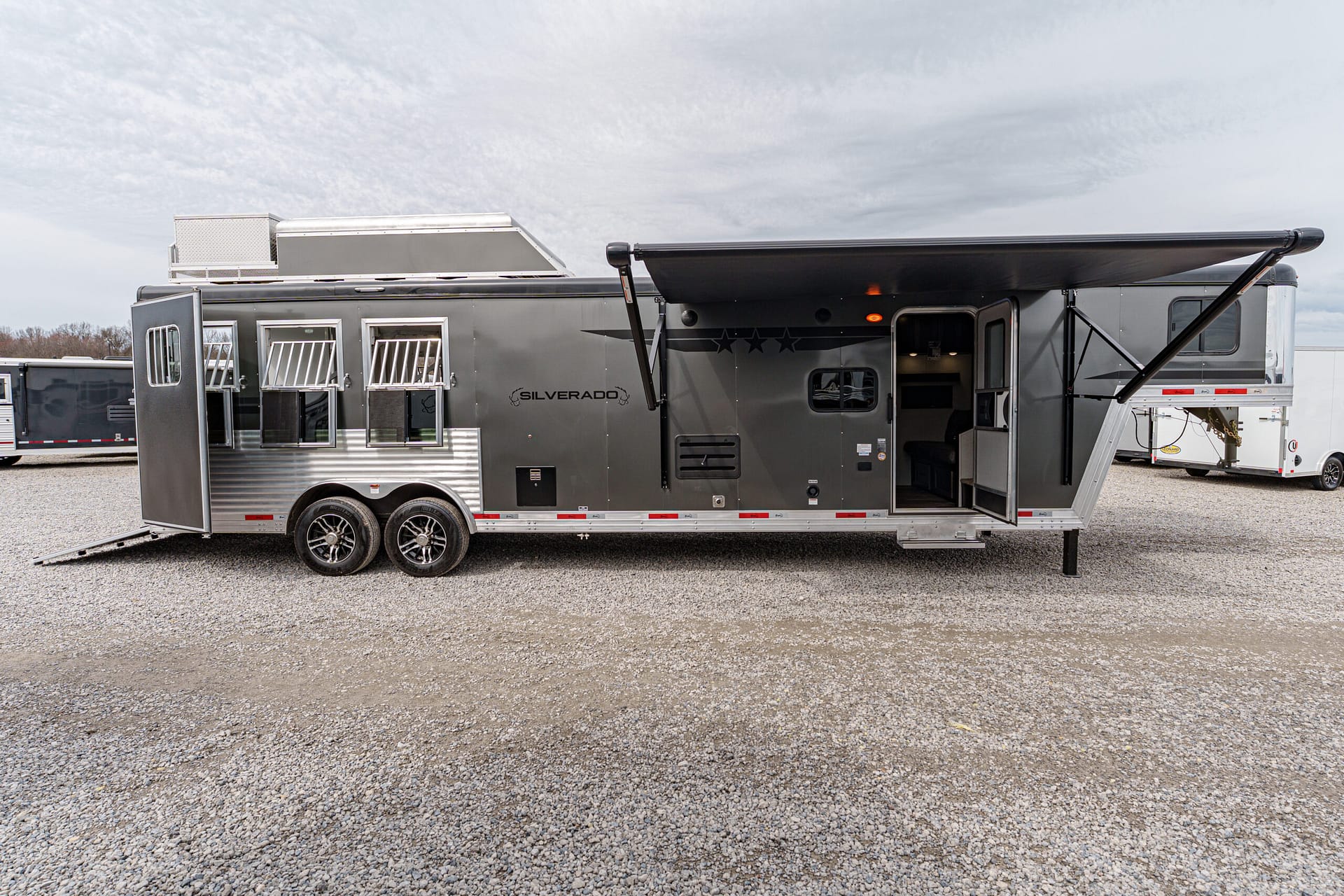 Grey travel trailer with extended awning, open entry door, and stairs on a gravel lot.