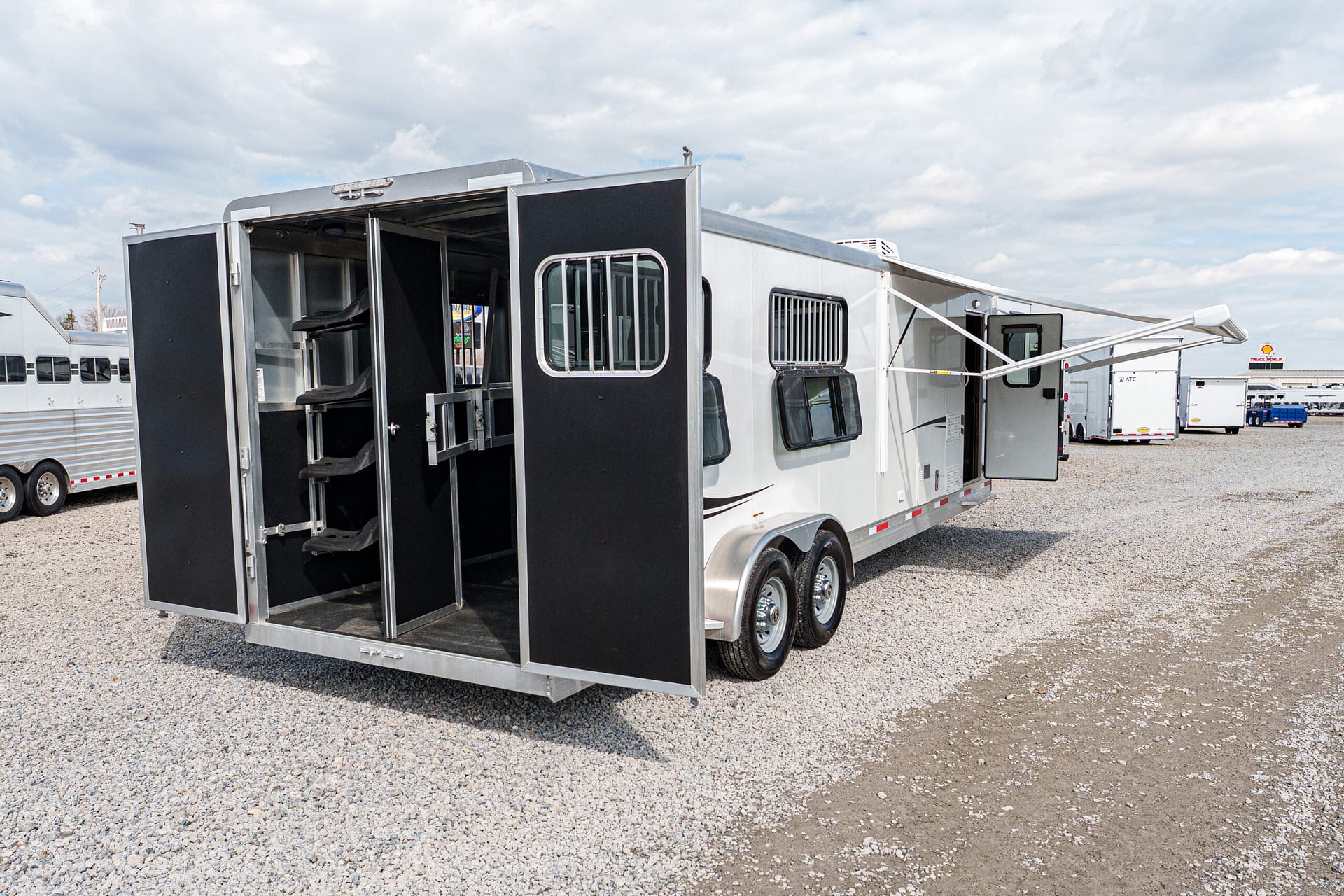 White horse trailer with the back doors open, showing interior stalls, parked on a gravel lot with other trailers in the background