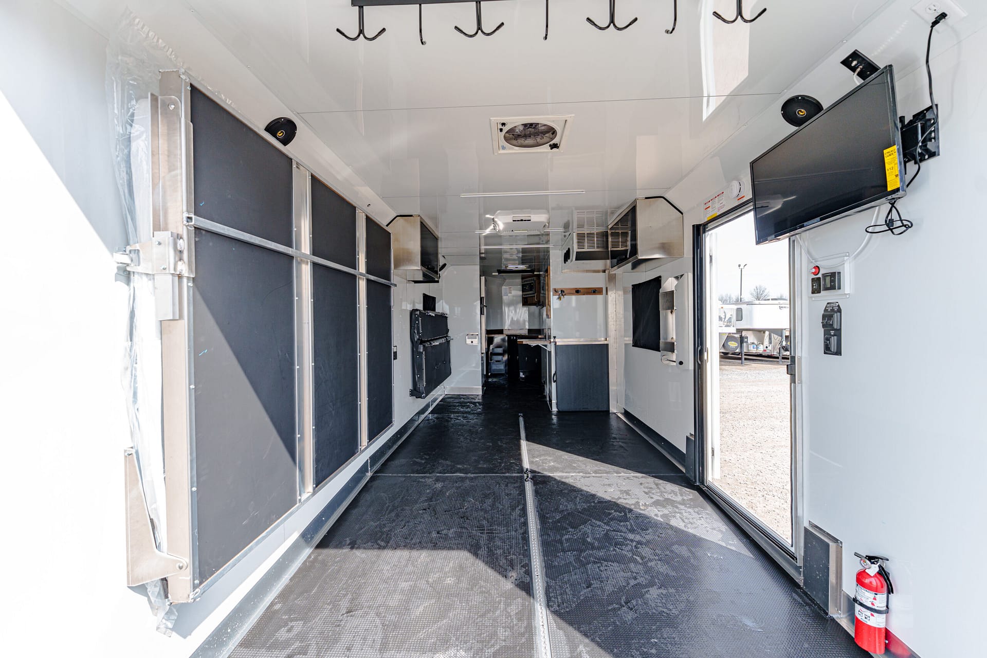 Interior of a white trailer with black storage panels along the left wall and a mounted flat-screen TV on the right, fire extinguisher near the entrance, and ceiling hooks overhead.