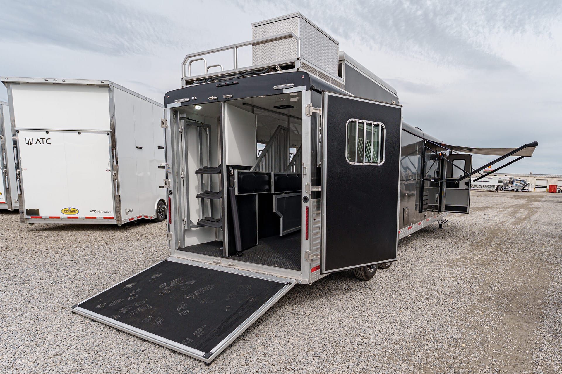 Open metal trailer with a lowered loading ramp on a gravel lot, interior stairs and seating visible inside; other trailers in the background.