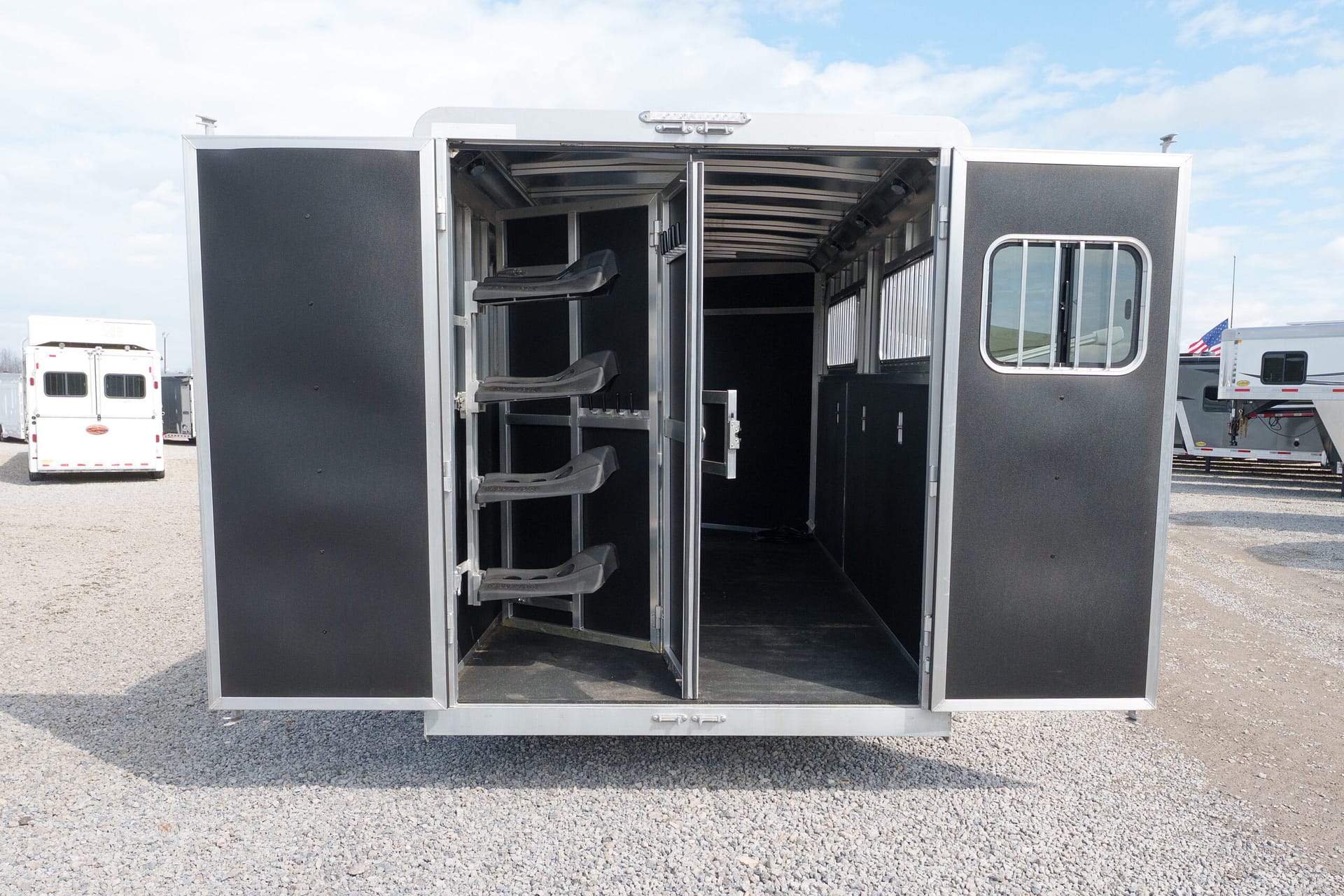 Open horse trailer interior with saddle racks along the left wall, exterior doors open, parked on gravel with other trailers in the background