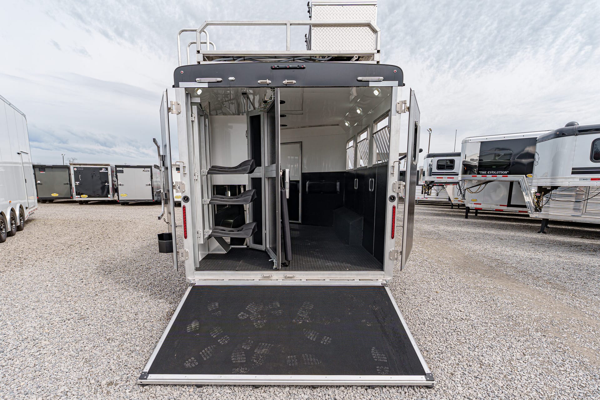 Open horse trailer with interior seating and a lowered ramp, parked among other trailers on a gravel lot.
