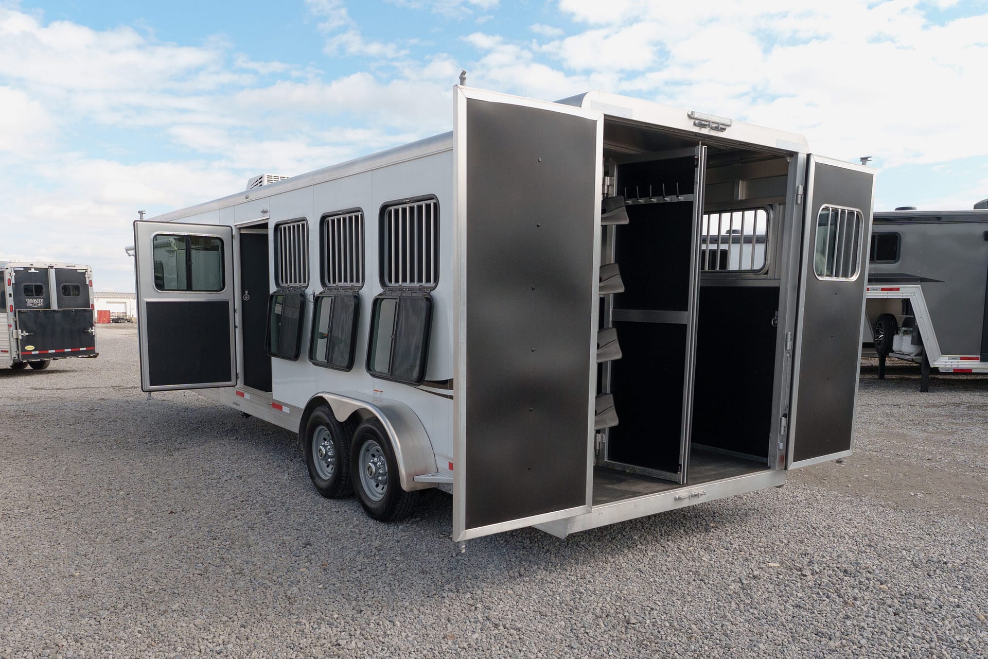 Silver horse trailer with multiple stalls and open side doors, parked on a gravel lot under a blue sky with clouds.