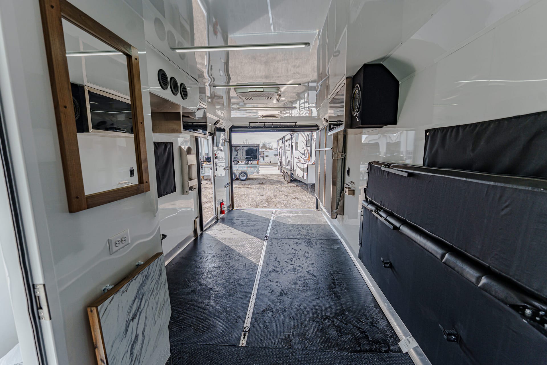 Interior of a glossy white RV hallway with black cushioned seating on the right and storage compartments; doorway at far end opens to an outdoor parking area.