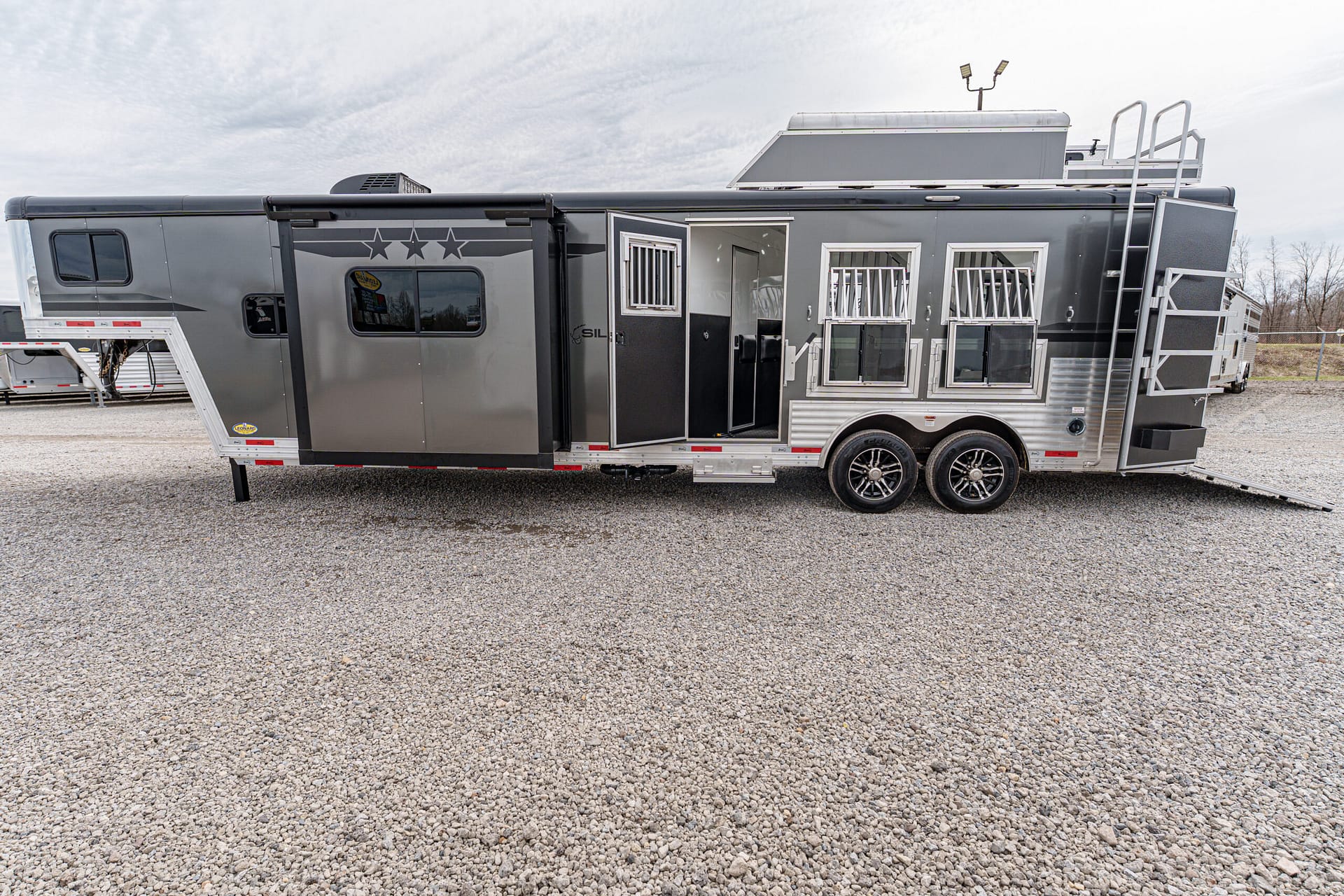 Long gray horse trailer parked on a gravel lot, showing side doors, windows with bars, and a rear ladder/ramp system.