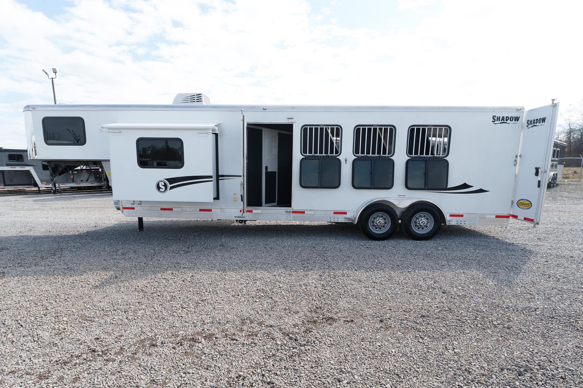 White horse trailer with barred windows and a small living area, parked on a gravel lot, Shadow logo on the side and front.