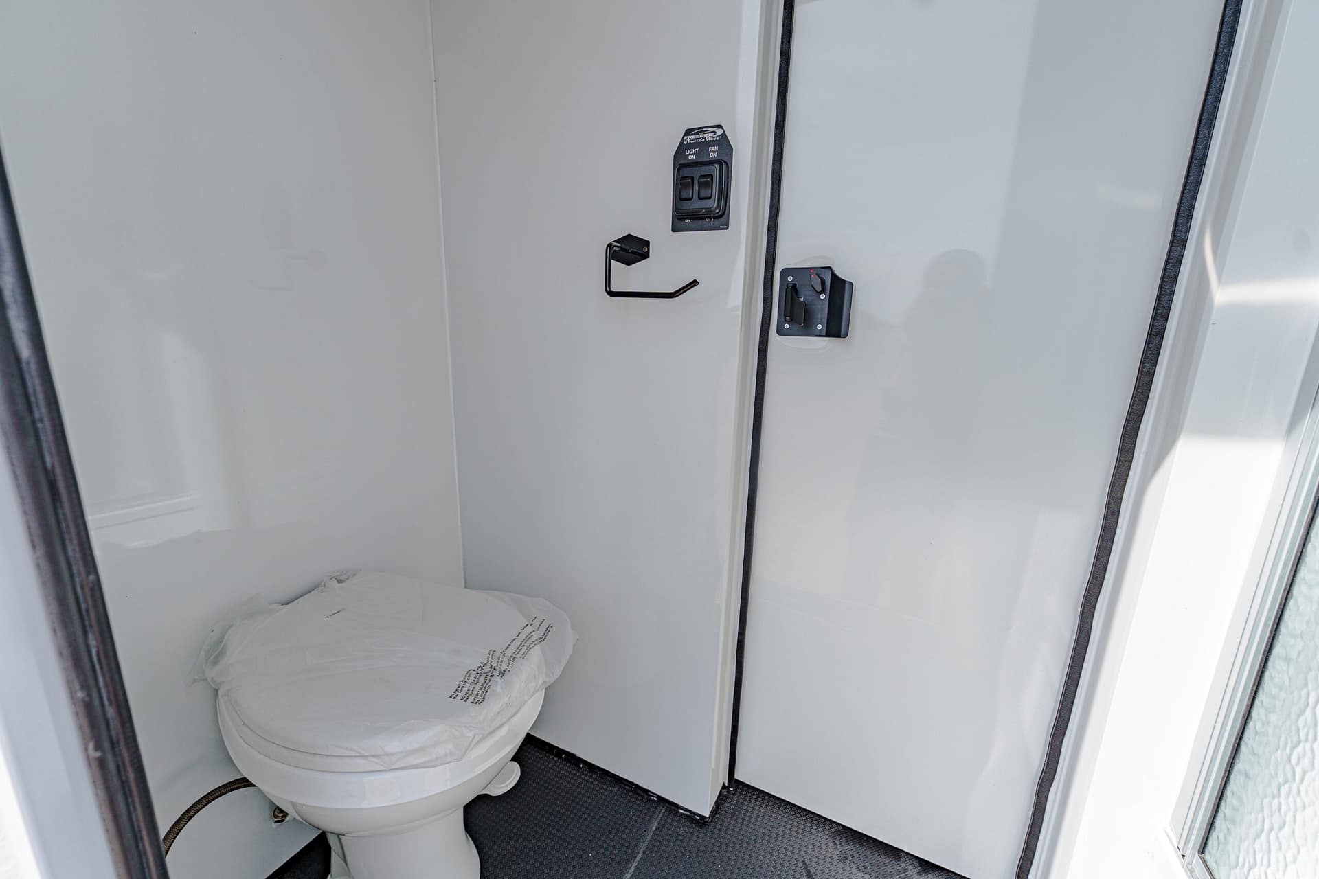 Small enclosed bathroom area with a white toilet, a black wall hook, and a control panel labeled light and fan on the wall beside the door.