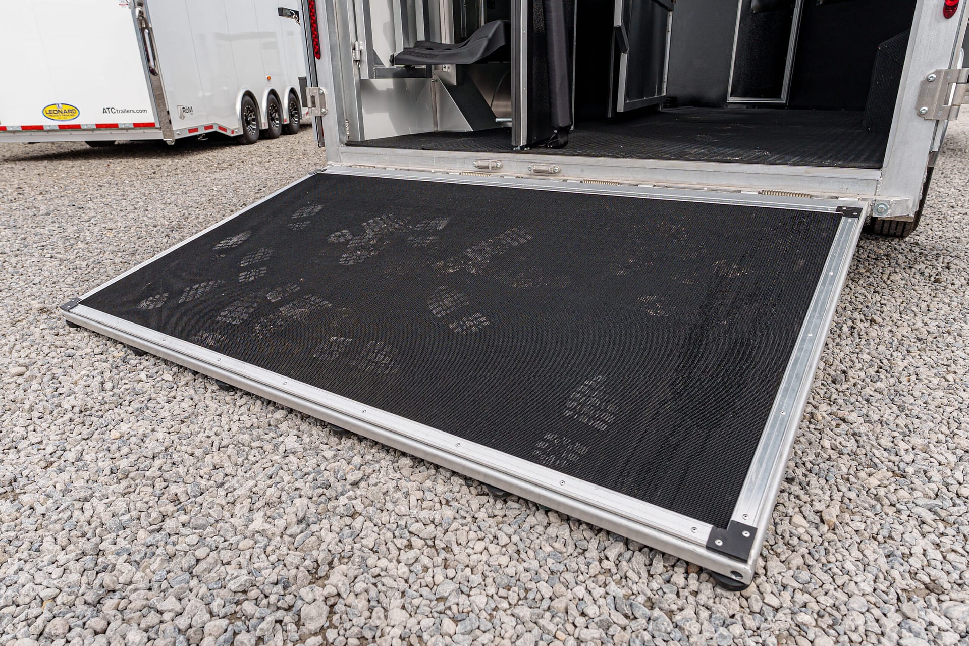 Open trailer ramp with a black textured non-slip surface, footprints visible, on a gravel lot with other trailers in the background.