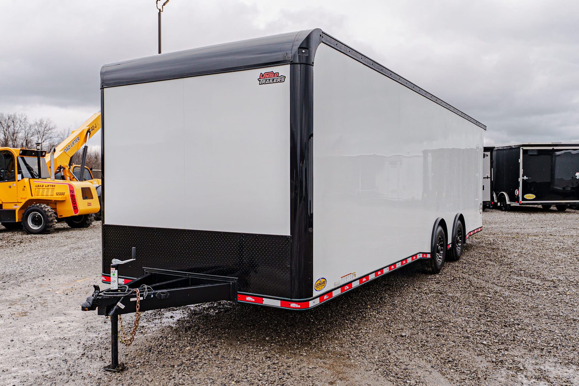 Dual-axle gray enclosed cargo trailer on a gravel lot, with a yellow forklift visible in the background.