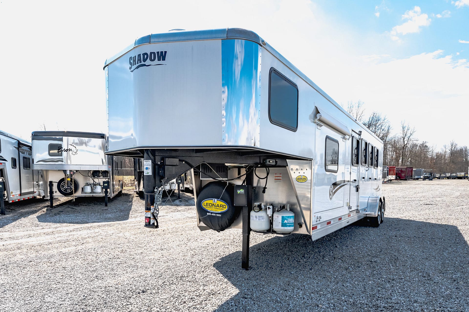 Silver fifth-wheel trailer with a Shadow logo parked on a gravel lot, with other trailers in the background and a blue sky above.