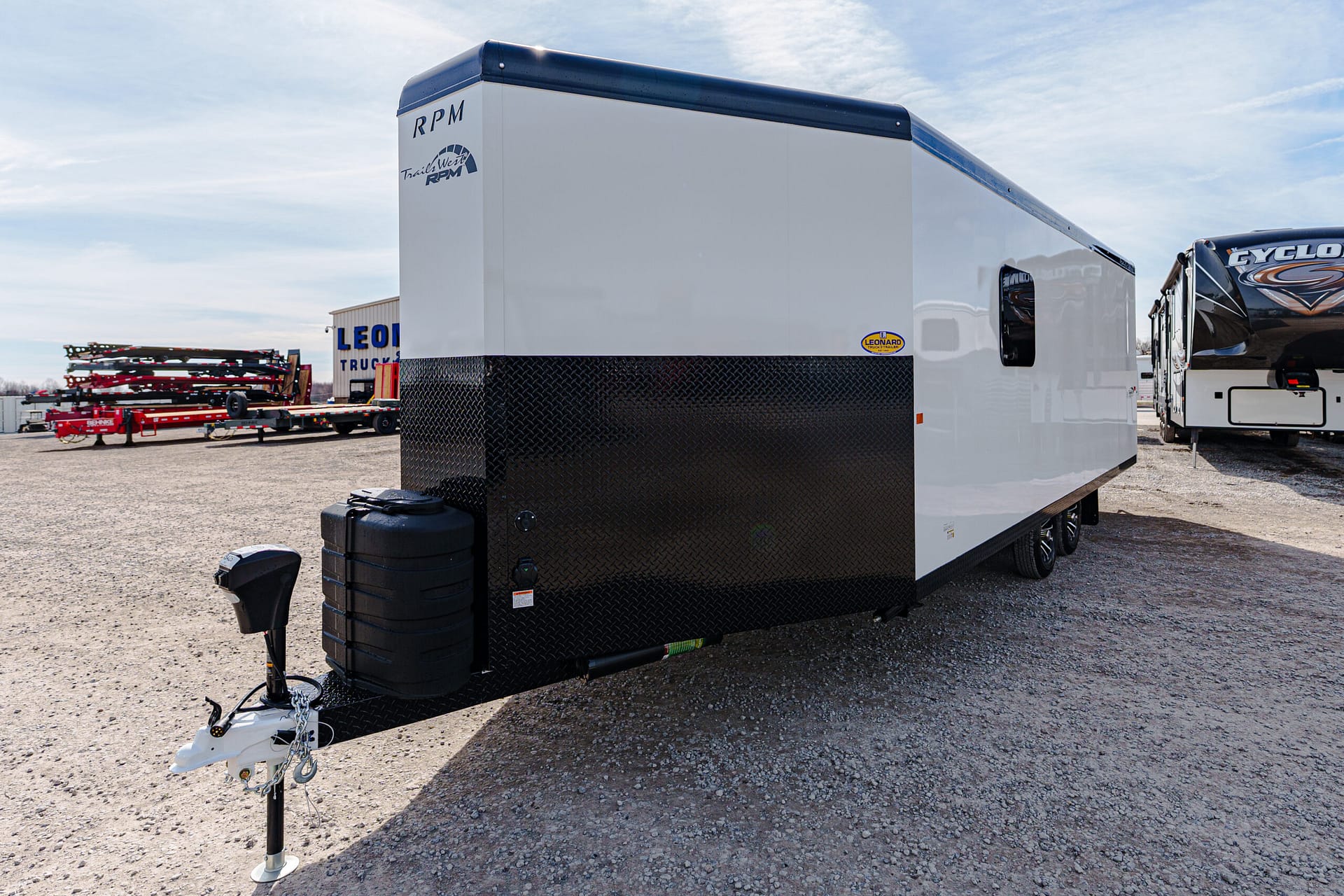 White travel trailer with a black diamond-plate front in a gravel lot, with other trailers nearby and a blue sky above at a trailer yard nowhere.