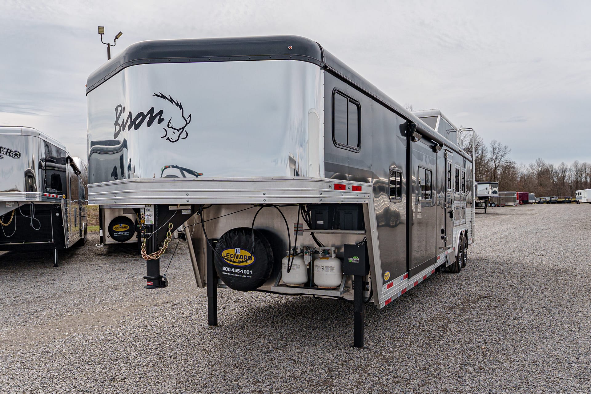 Front-right view of a glossy silver fifth-wheel trailer raised on leveling jacks in a gravel lot, with propane tanks and a spare tire cover visible at the front edge.