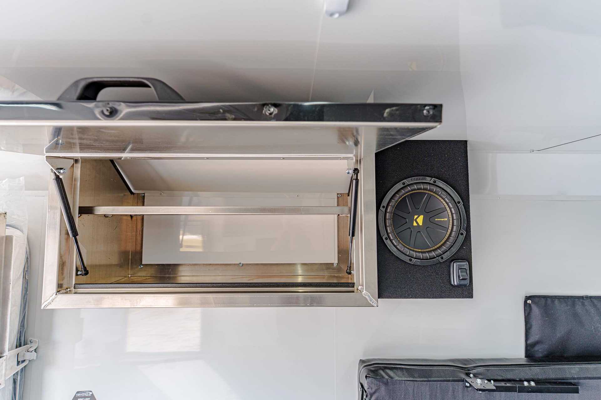 Open stainless steel service hatch in a trailer kitchen, with metal shelving and a large round speaker mounted on a black wall panel to the right.