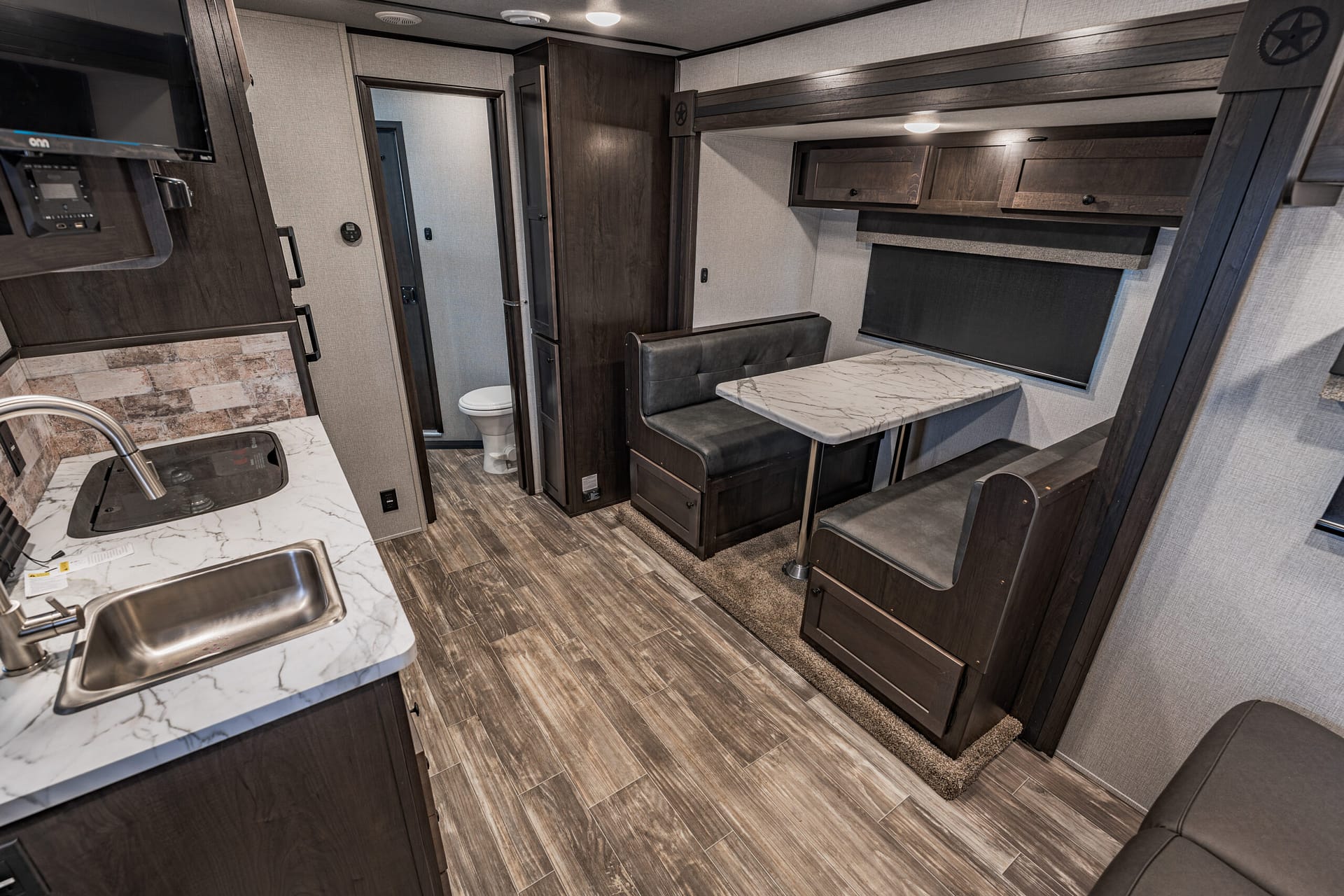 Interior of a modern RV with a kitchen island sink, marble countertop, and dark wood cabinets; there is a dining booth with a marble-top table and padded seating opposite a window with a shade, plus overhead storage and a hallway to a bathroom door in the back.
