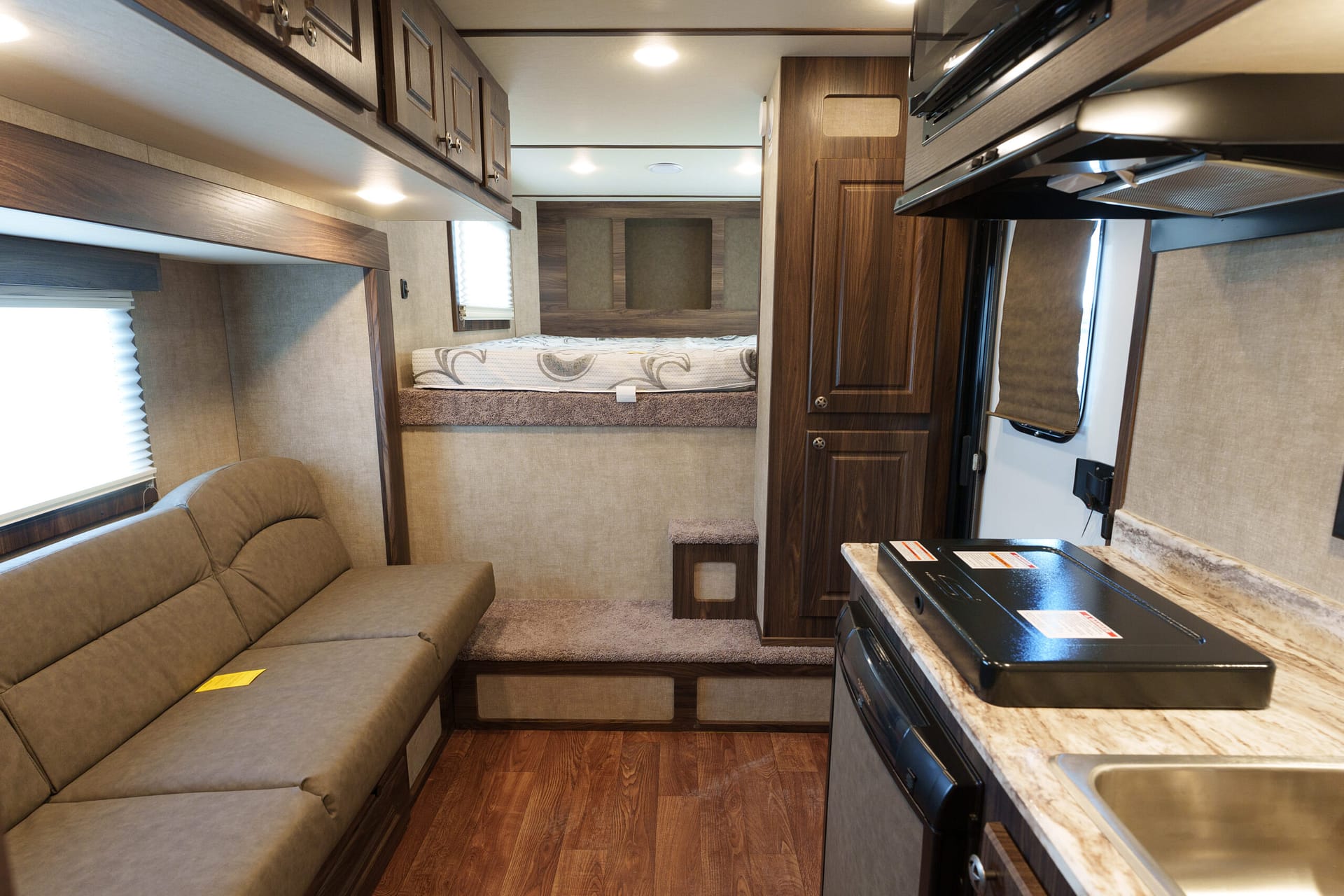 Interior of a cozy RV: sofa on left, kitchen counter and stove on right, bed at back with wood paneling and windows nearby.