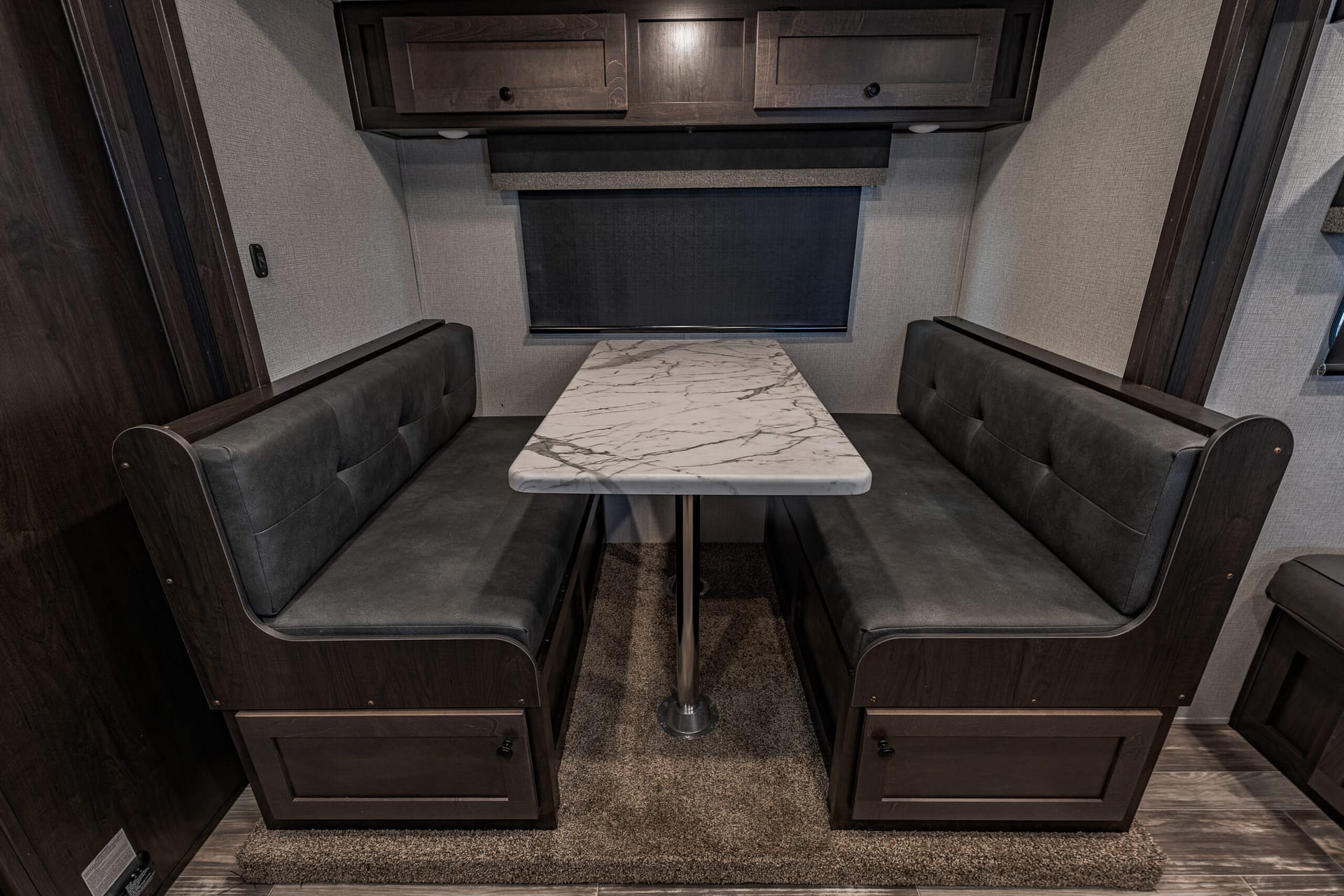 RV dinette with dark wood cabinets, marble tabletop, and grey upholstered bench seating around a windowed wall.