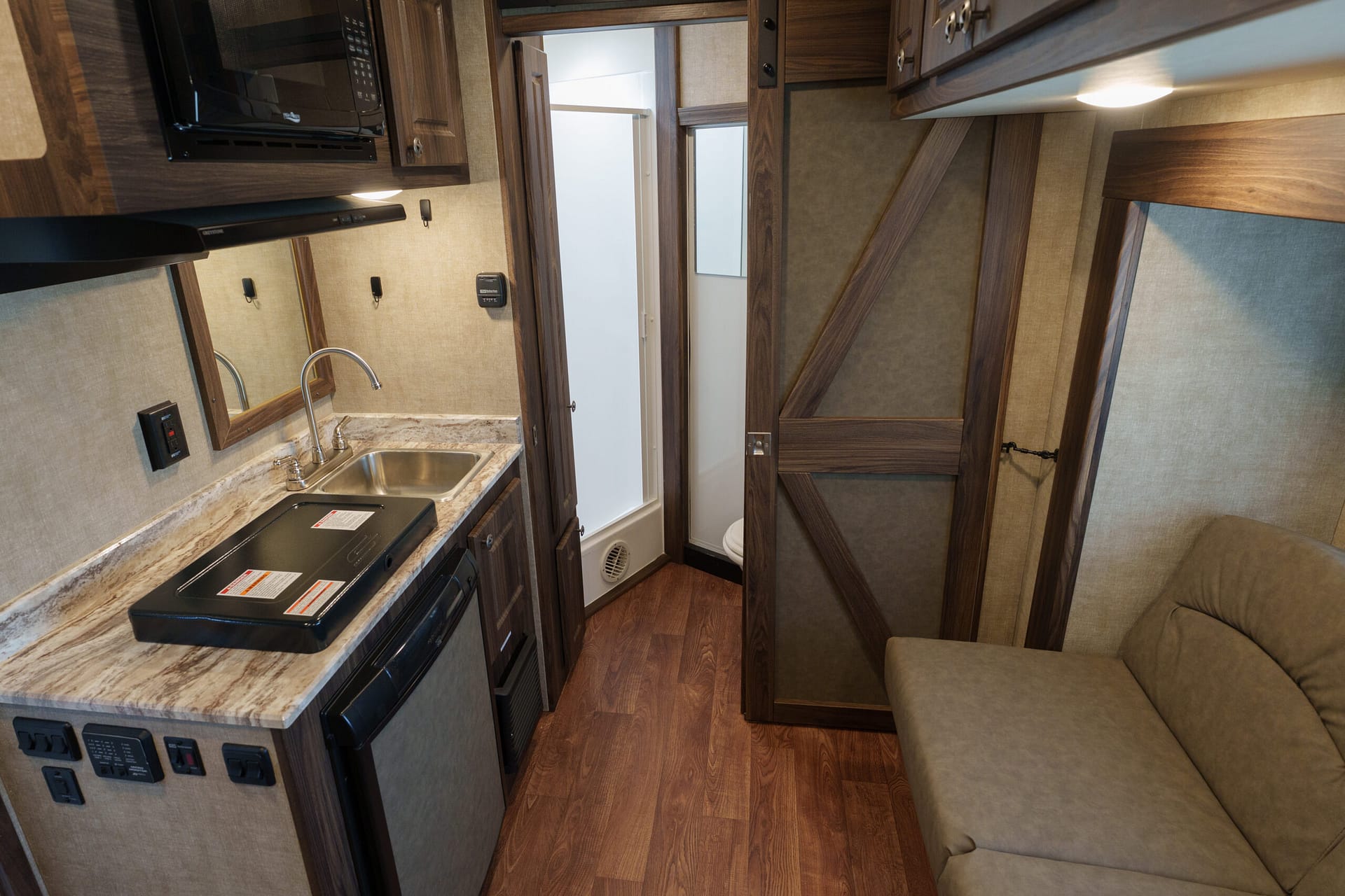 Compact RV kitchen with laminate counter, stainless sink, and built-in appliances amid dark wood cabinets and a beige sofa.