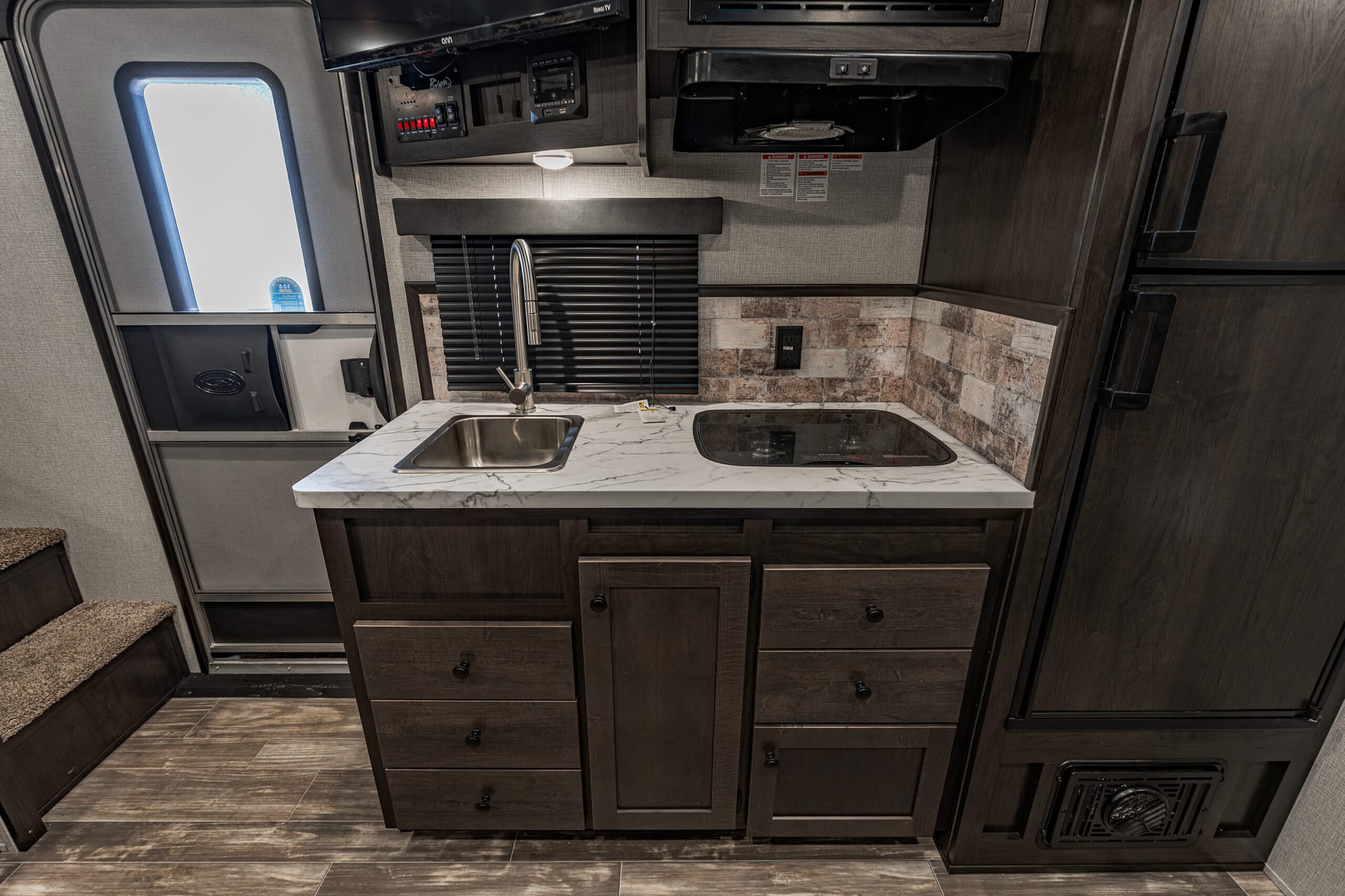 Rv kitchen interior with dark wood cabinets, marble countertop, stainless sink and faucet, stove, and window blinds