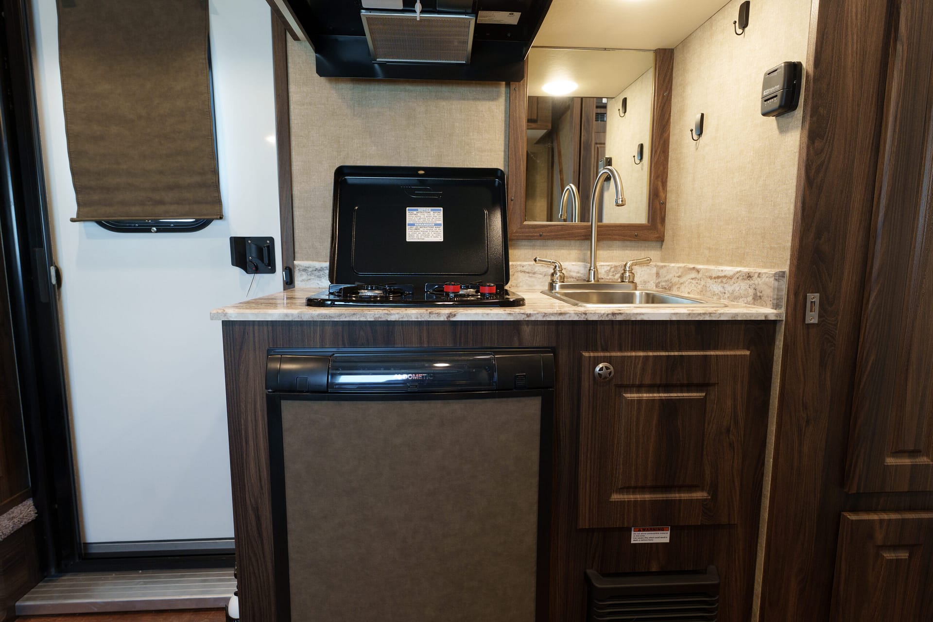 RV kitchenette with open two-burner stove, stainless sink, tall curved faucet, and dark wood cabinets along beige walls. A window shade is on the left.