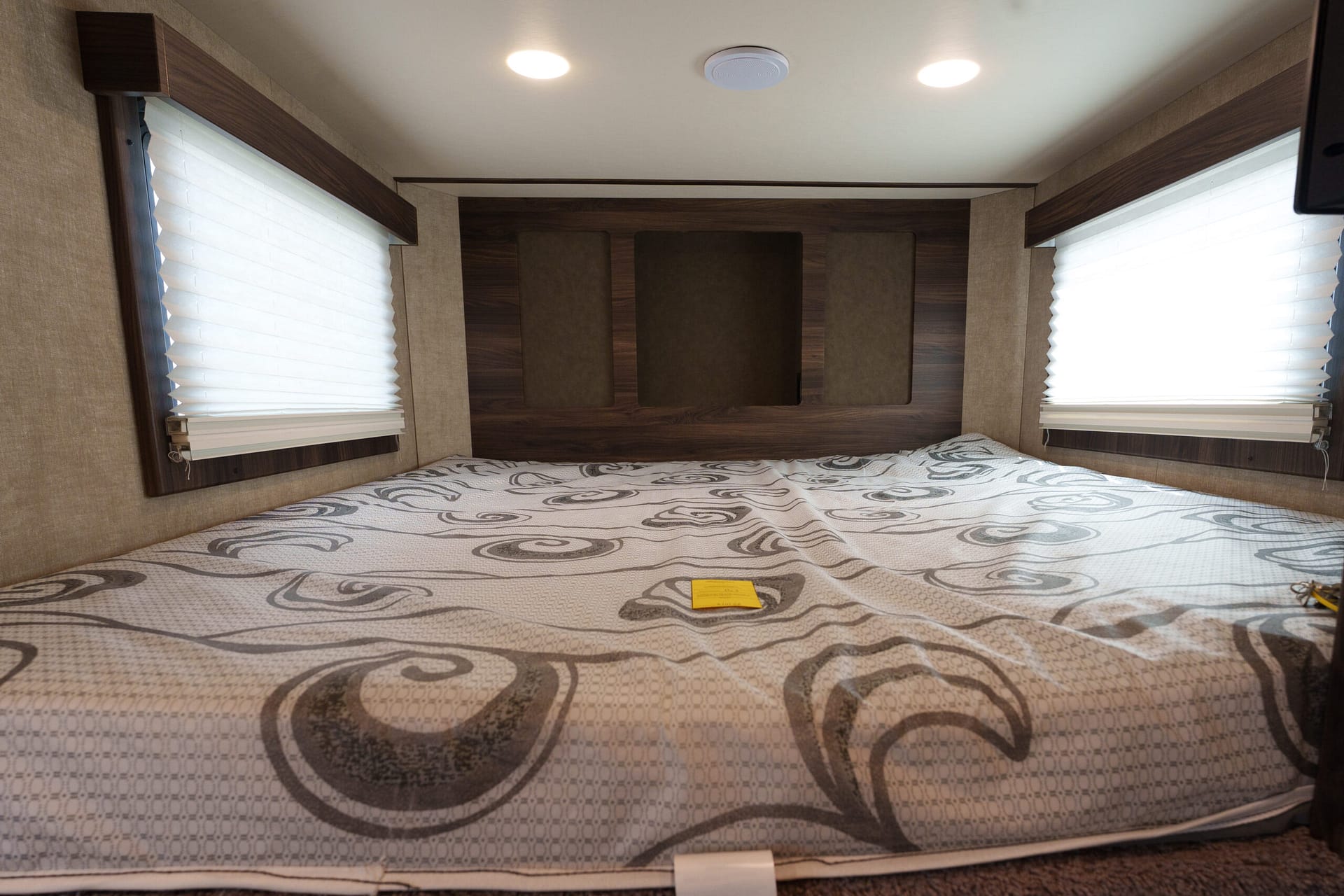 RV bedroom with a patterned quilt, dark wood headboard, and two windows with white blinds.