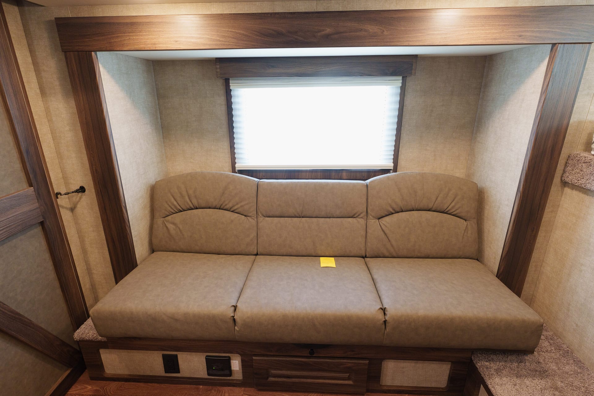 Beige upholstered sofa bed with wood framing in a small RV bedroom, centered by a window with white blinds behind it.