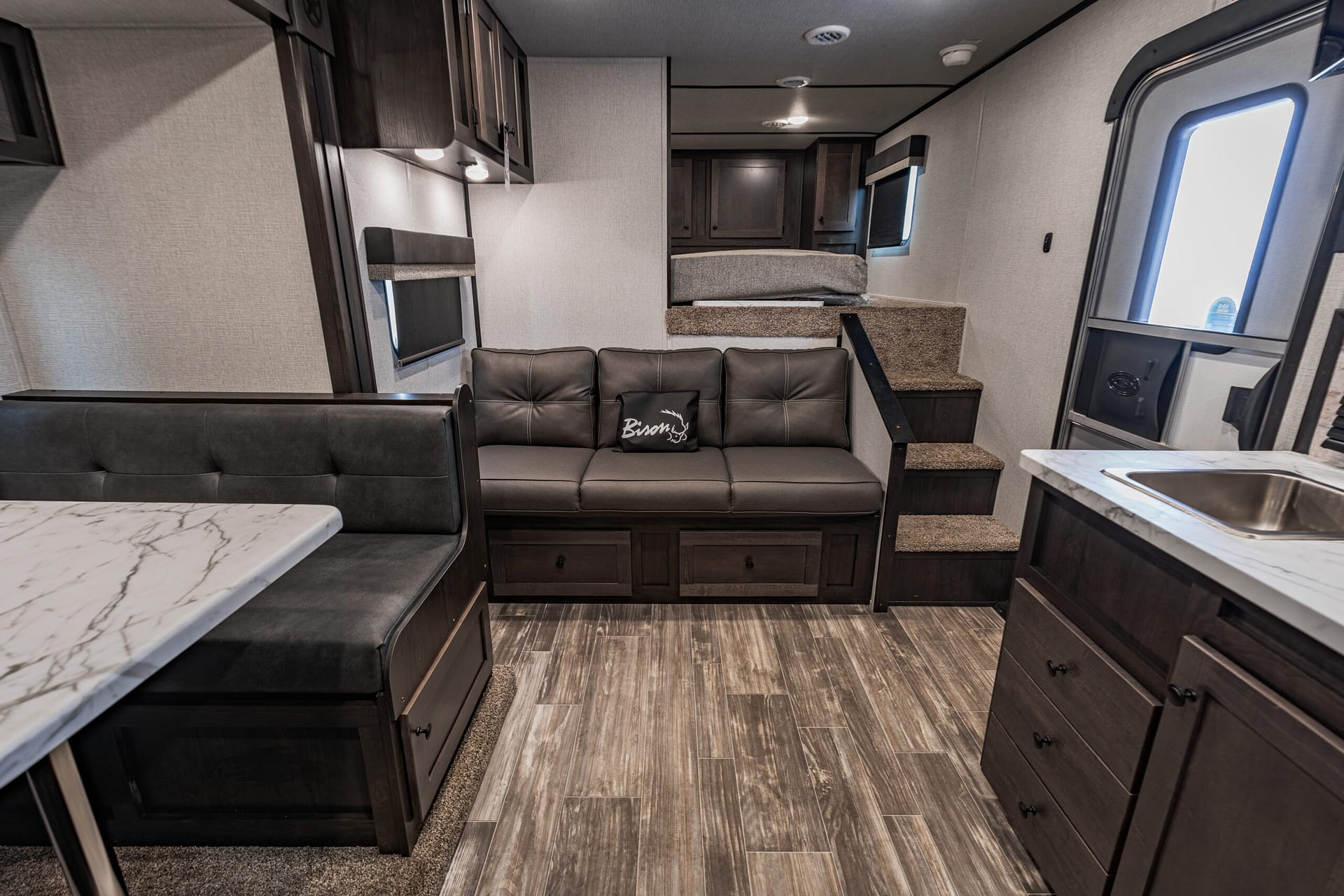 Interior view of a modern RV living area with a dark leather sofa, stairs to a loft, and overhead cabinets.