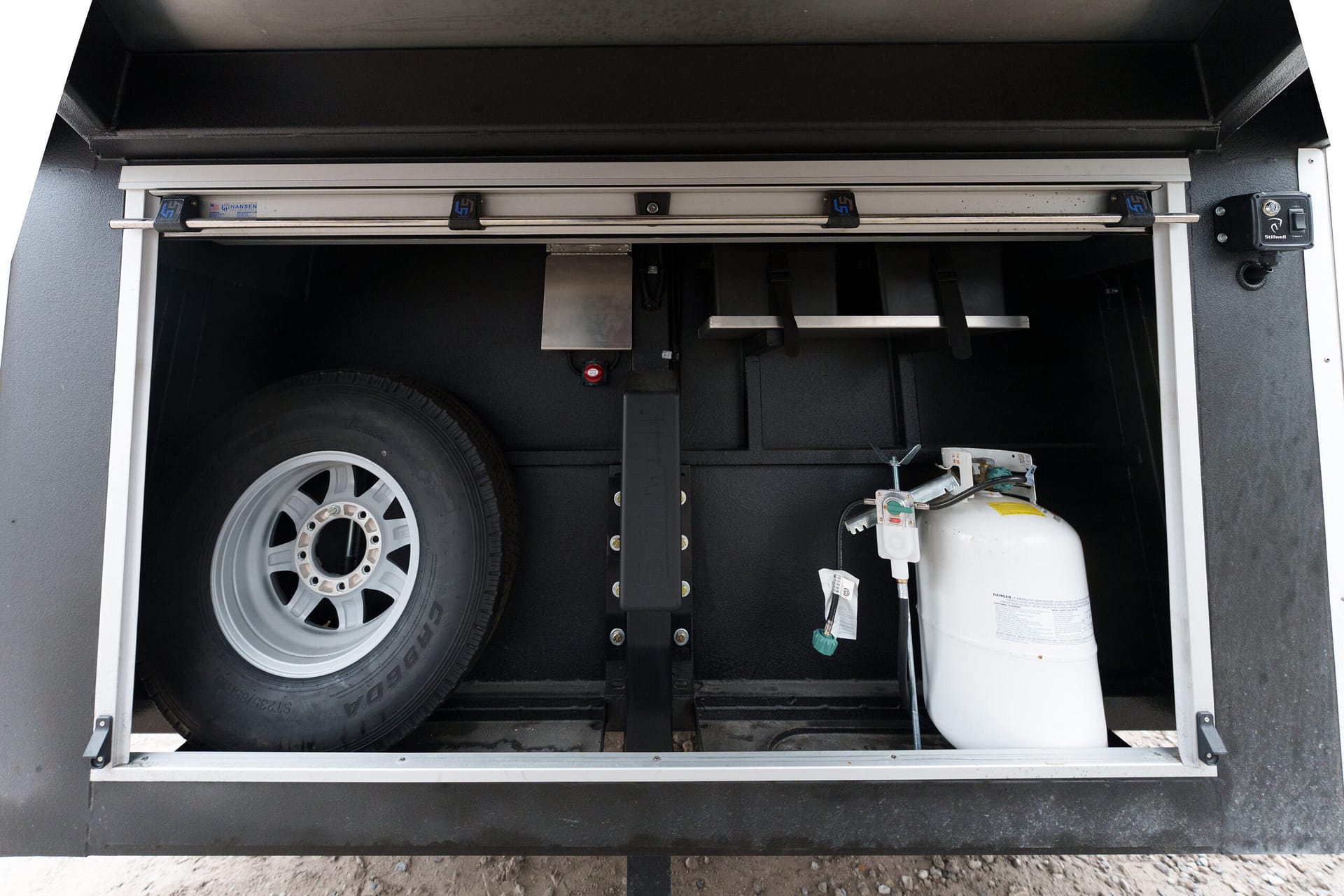 Interior of a utility trailer storage area showing a spare wheel on the left, a white propane tank on the right, and metal mounting rails across the top.