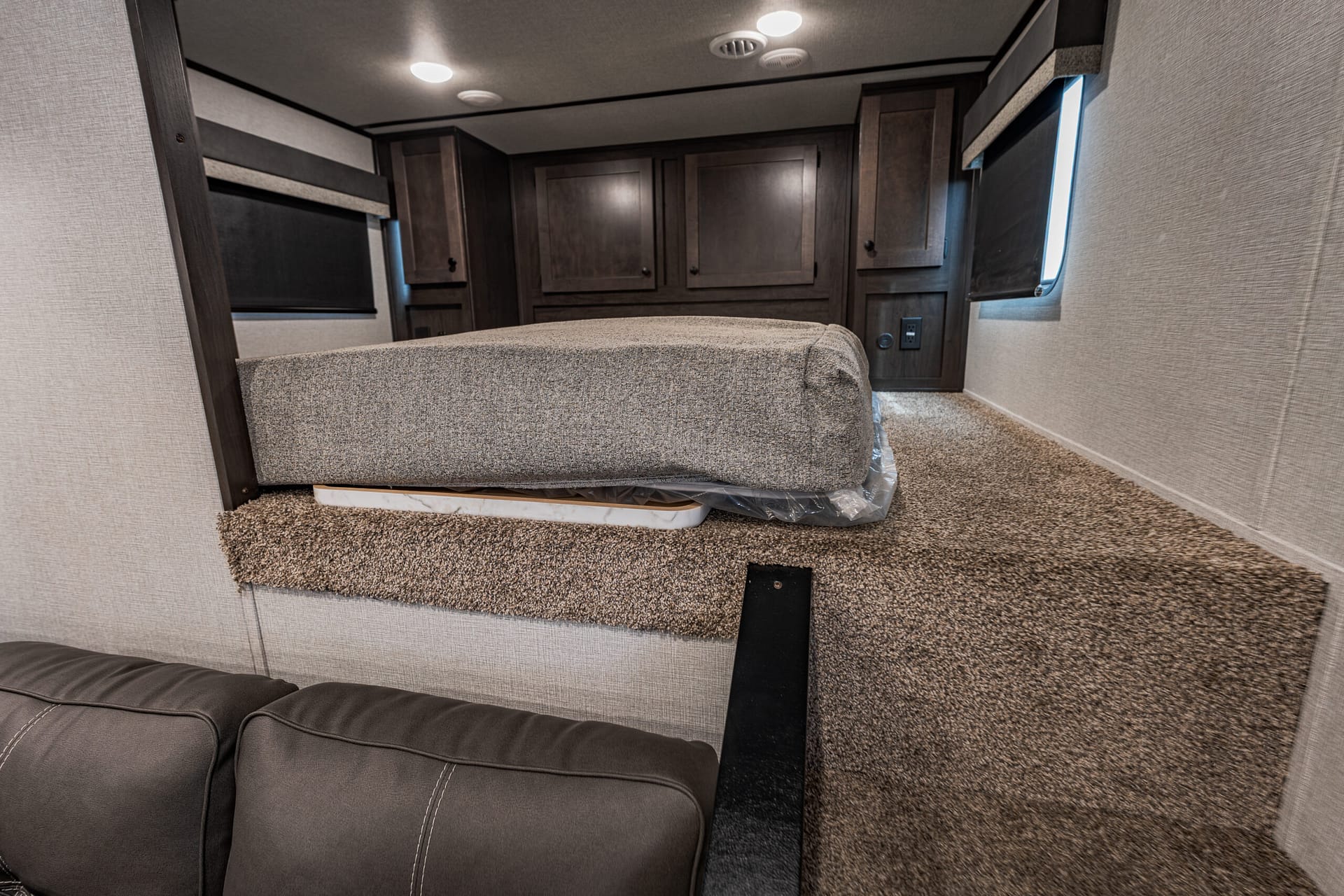 RV bedroom interior with a padded bed, dark wood cabinets, and carpeted steps in the foreground.