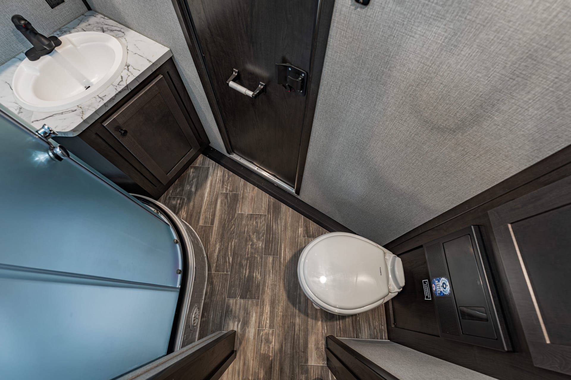 Compact RV bathroom with a marble countertop sink, blue curved shower enclosure, dark wood cabinets, and a toilet.