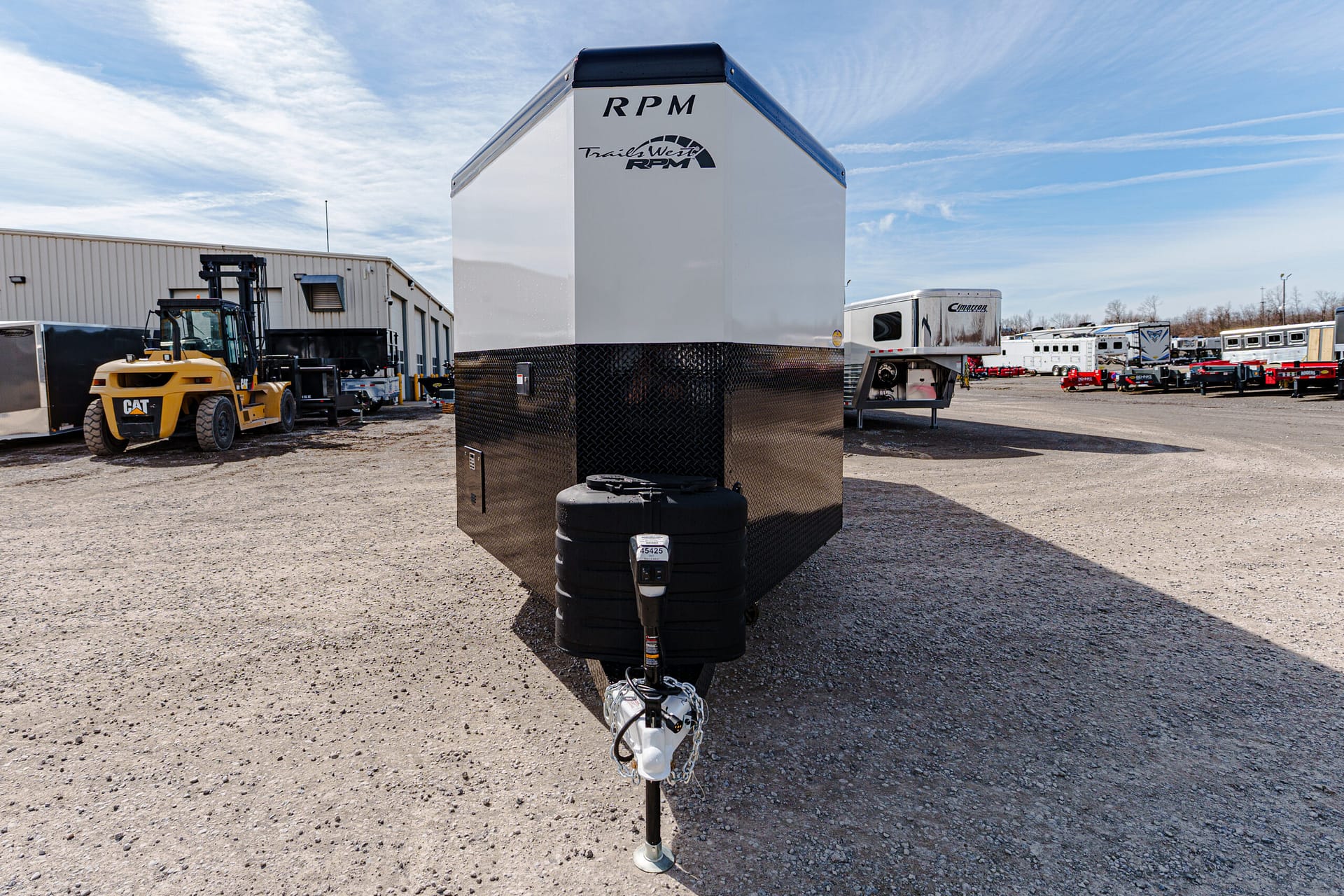 Front view of a tall white RPM trailer with a black diamond-plate base, in a gravel trailer yard with forklifts and other trailers nearby.