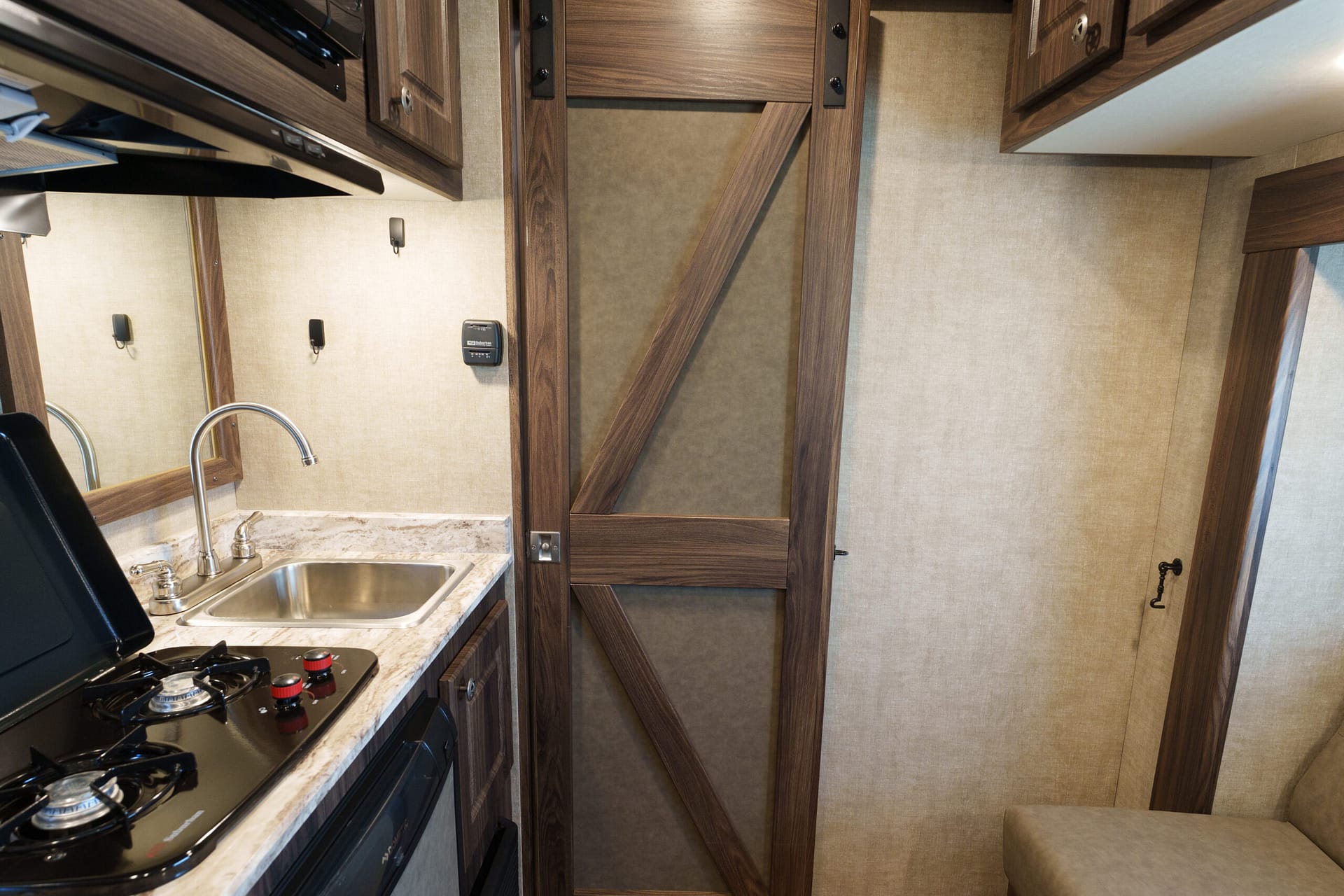 Compact RV kitchen with stainless sink, marble countertop, and gas stovetop on the left, surrounded by dark wood cabinets.