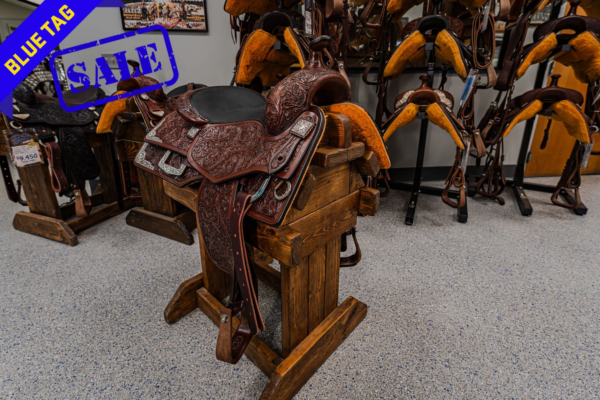 Ornate brown western saddle on a wooden stand in a tack shop, with a blue sale tag across the top left.