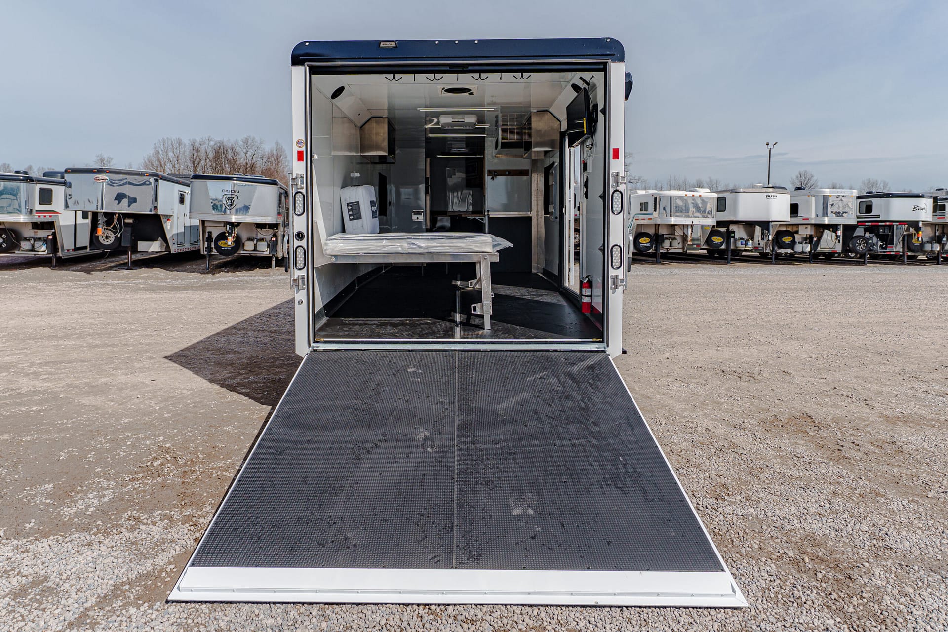 Open rear of a white mobile kitchen trailer with a metal ramp extended, showing stainless steel appliances inside and other trailers in a gravel lot behind.