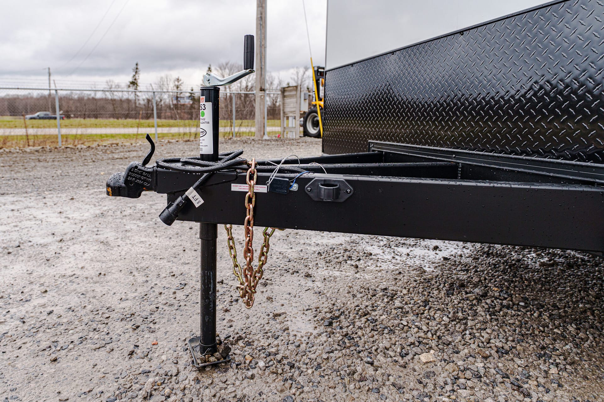 Close-up of a trailer tongue with a vertical jack, safety chains, and a diamond-plate trailer wall in the background on a gravel lot.