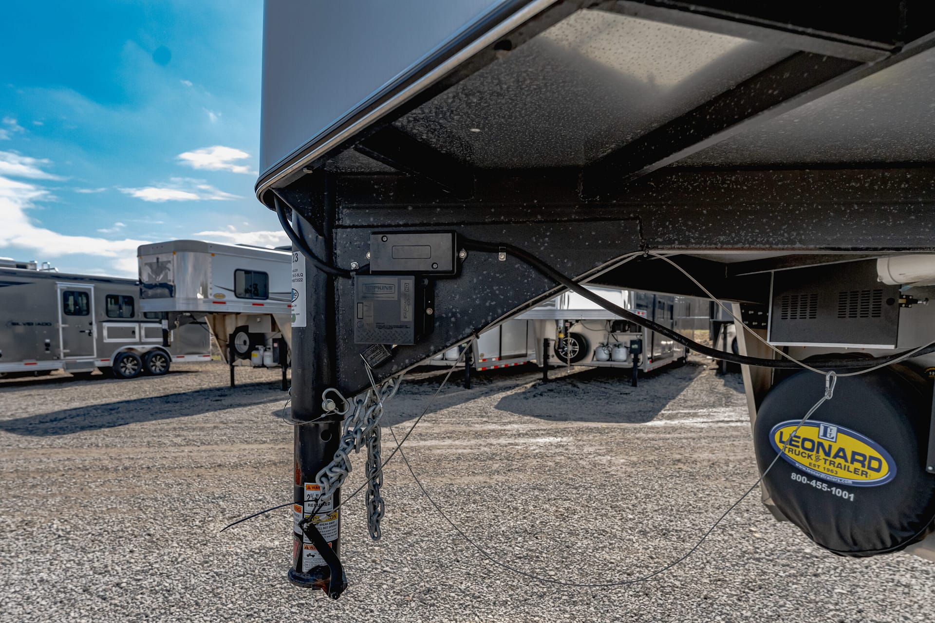 Close-up of a fifth-wheel trailer hitch with safety chains and cables, in a gravel lot with other RVs in the background under a blue sky.
