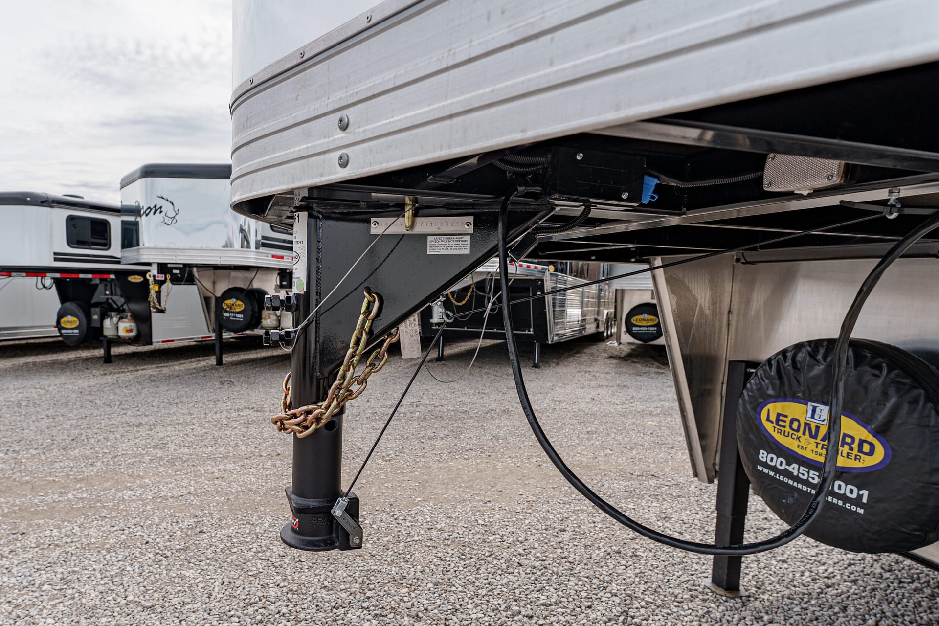 Close-up of a trailer hitch area with chains, wiring, and support legs under a silver RV trailer on gravel ground (informational).