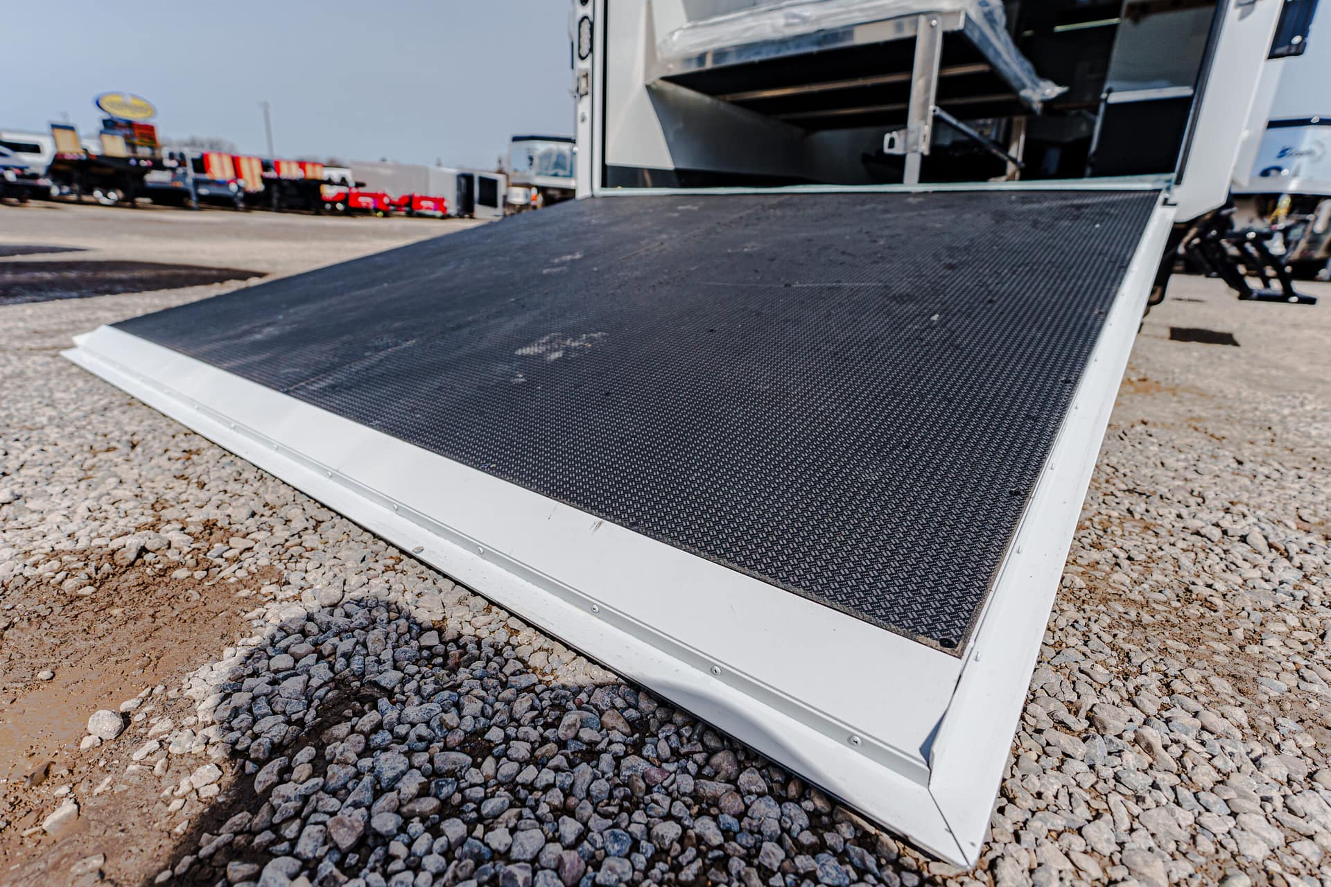 Close-up of a black textured loading ramp attached to a white truck in a gravel lot with other trucks in the background.