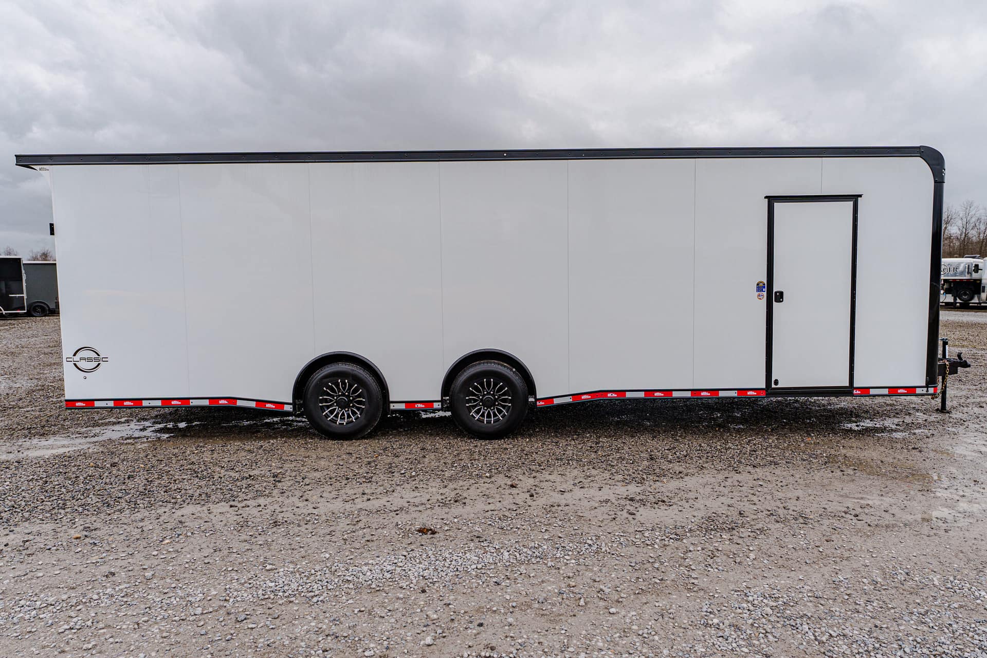White enclosed cargo trailer with a side door, parked on a gravel lot under a cloudy sky.