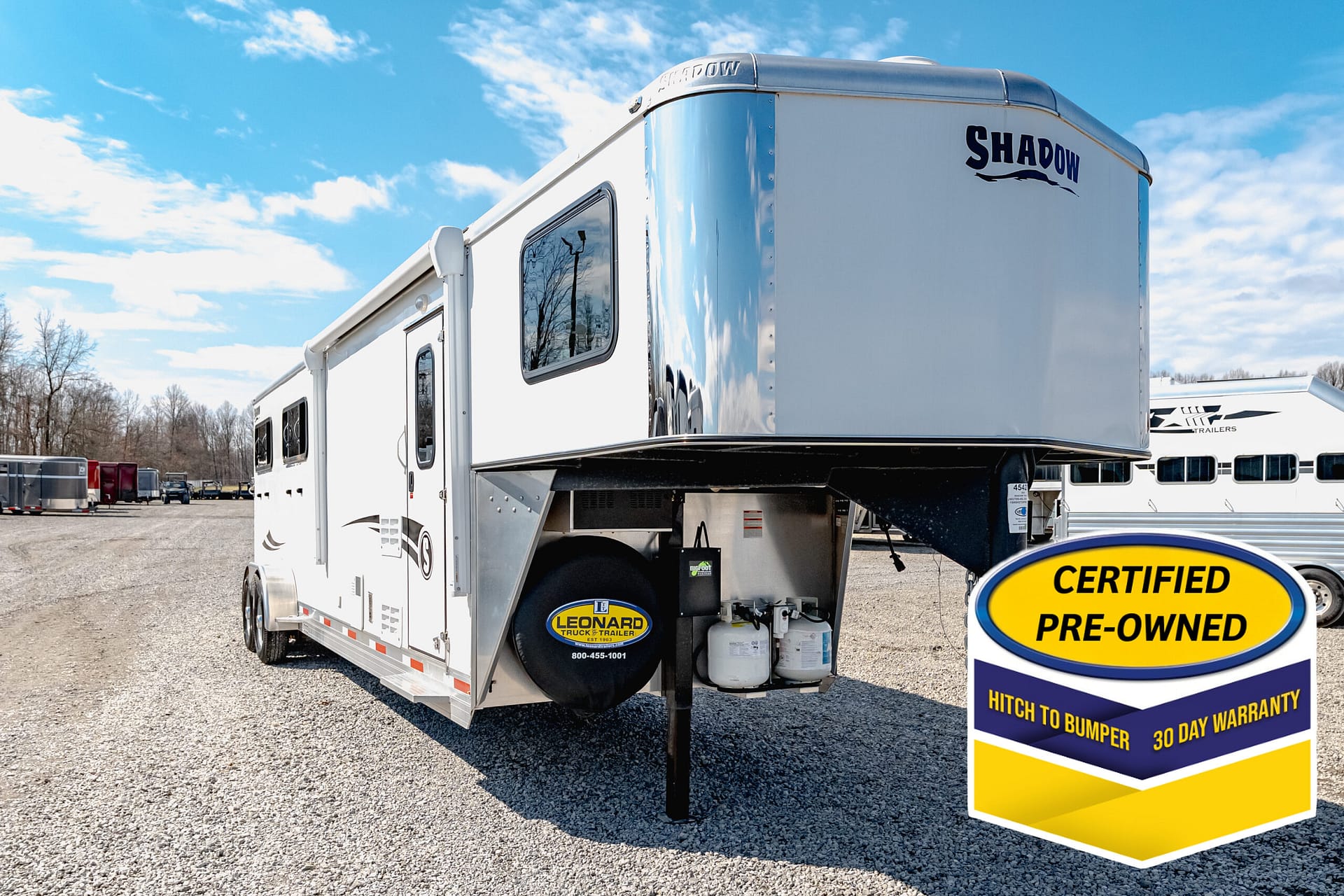 White gooseneck trailer with a 'Shadow' logo, parked on gravel under a blue sky; a large yellow 'CERTIFIED PRE-OWNED' sign is in the foreground.