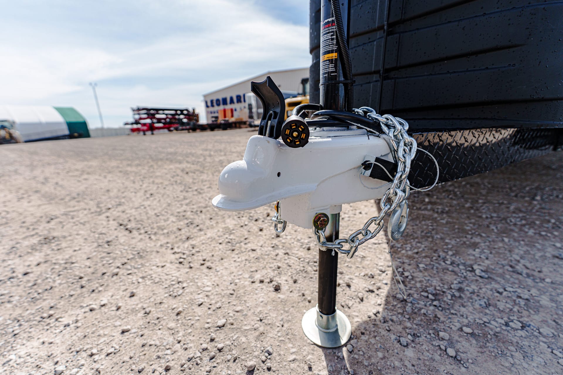 Close-up of a trailer hitch with safety chain and winch on a gravel lot, equipment blurred in the background.