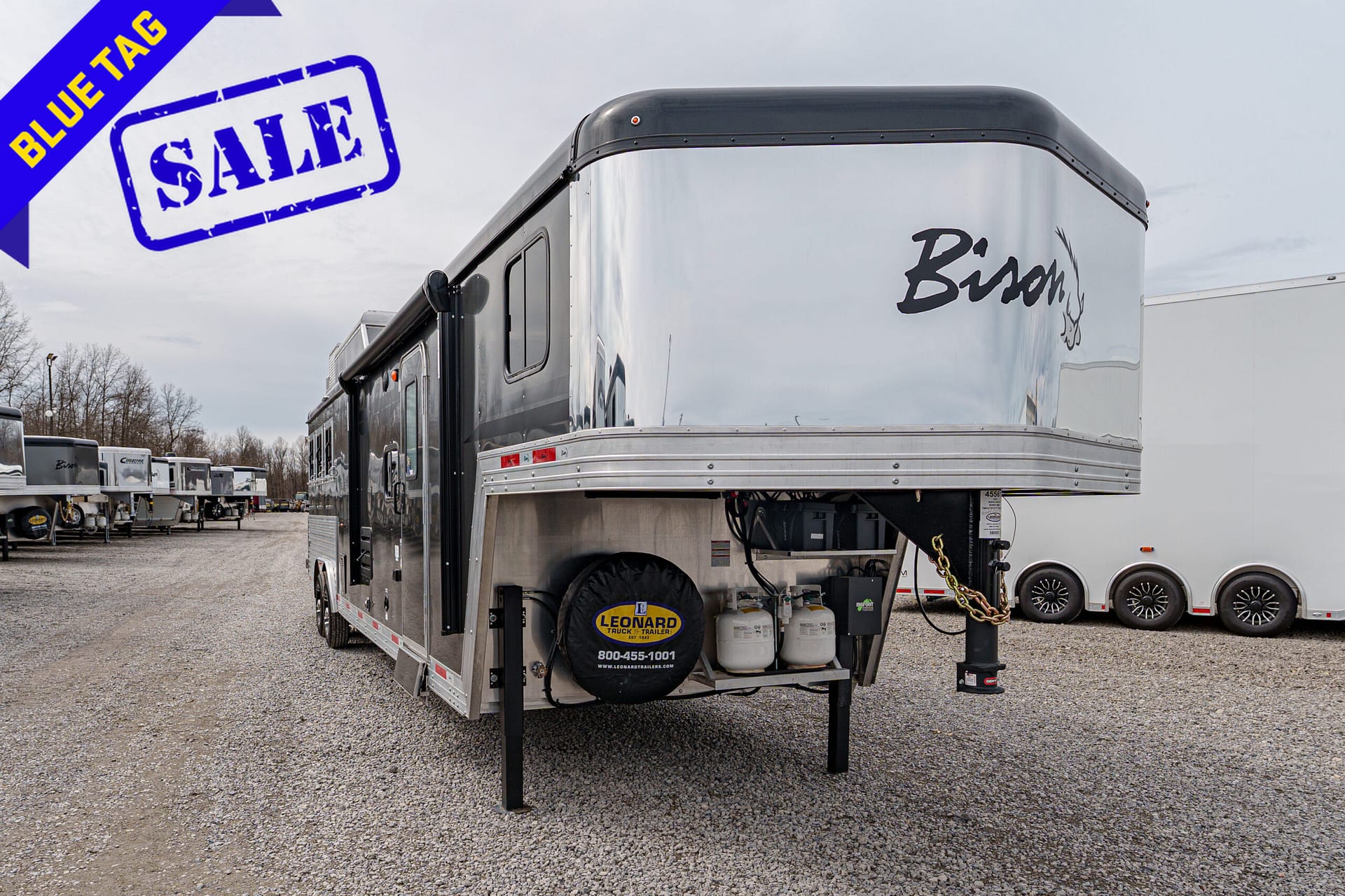 Silver horse trailer on a gravel lot with a large blue 'SALE' stamp and a diagonal blue ribbon reading 'BLUE TAG' in the top-left corner.